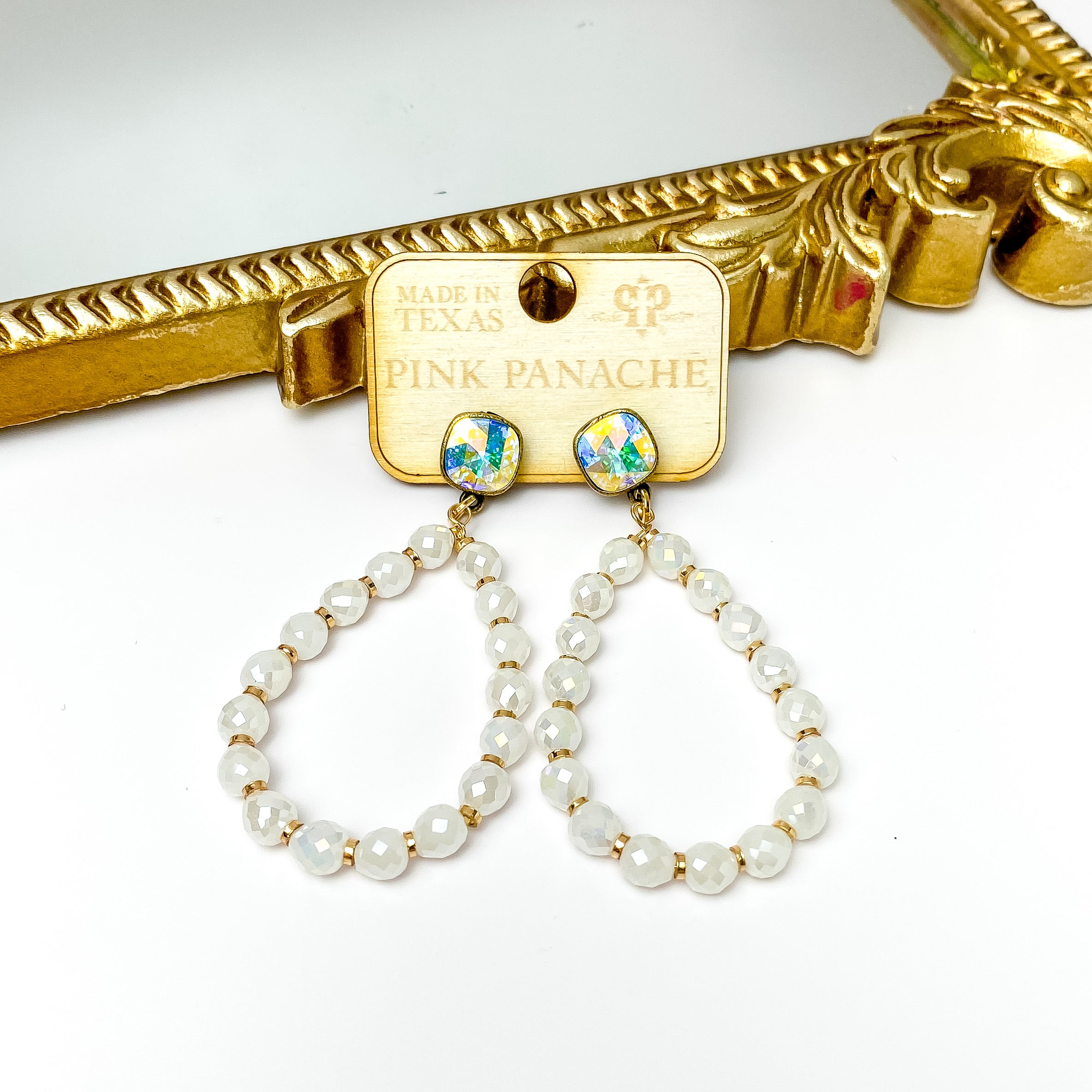 AB cushion cut post back earrings with a teardrop pendant. This pendant includes white crystal beads ith small gold spacers. These earrings are pictured on a wood earrings holder in front of a gold mirror on a white background. 