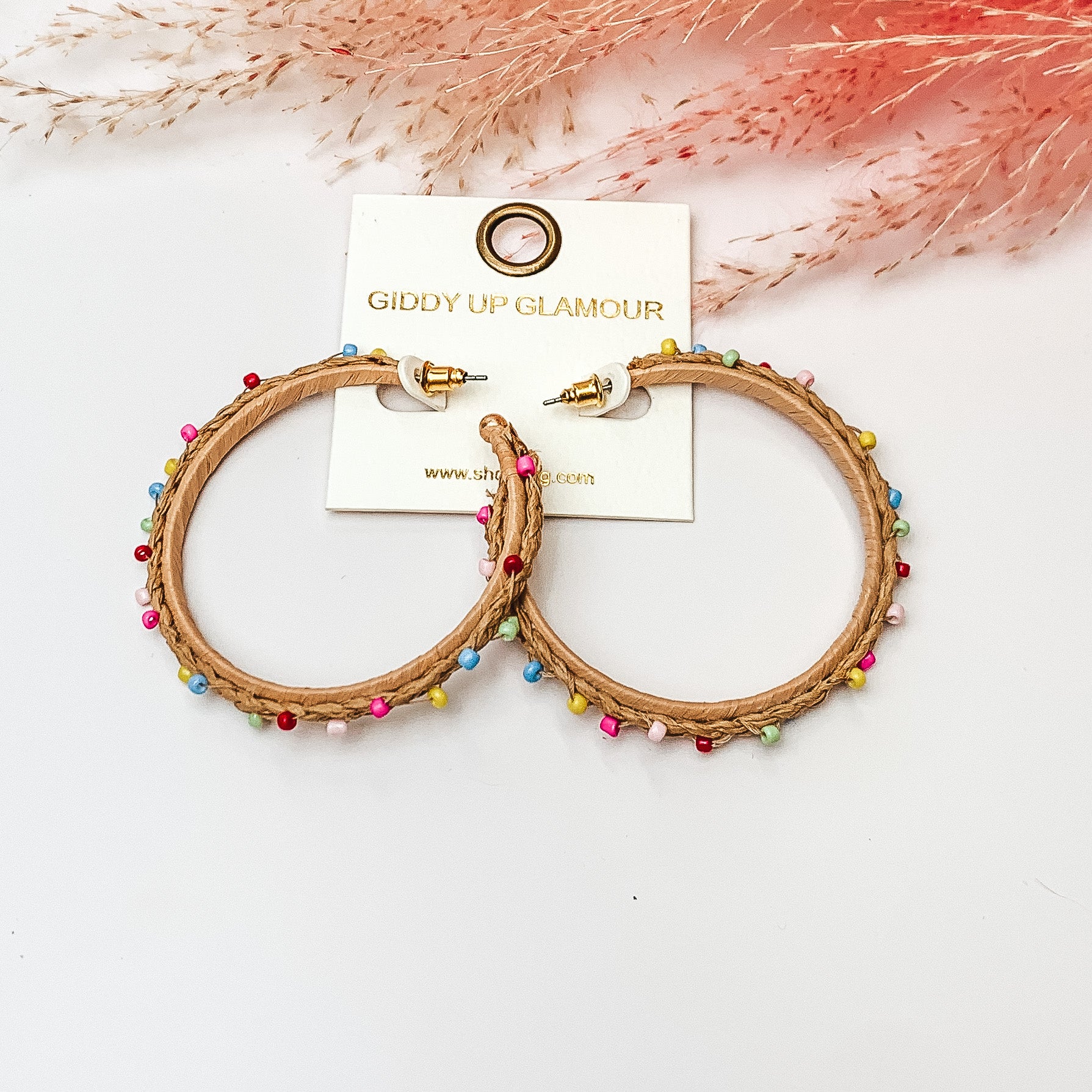 Pictured are raffia braided hoop earrings in tan with colorful beads. They are pictured with a pink feather on a white background.
