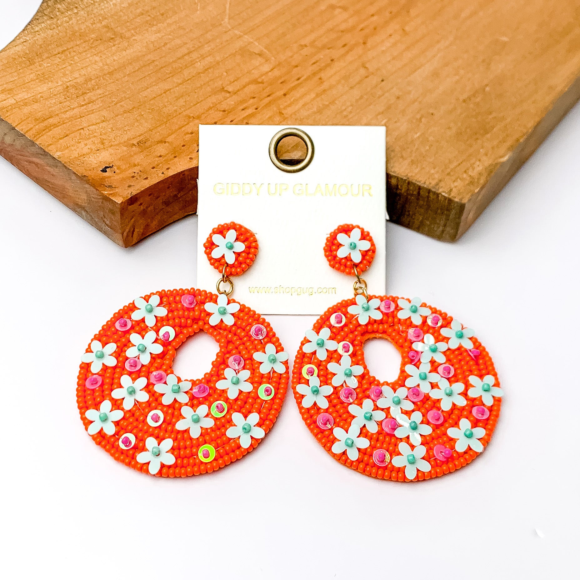 Orange beaded circular drop earrings with pink and blue designs. Pictured on a white background with a wood piece at the top.