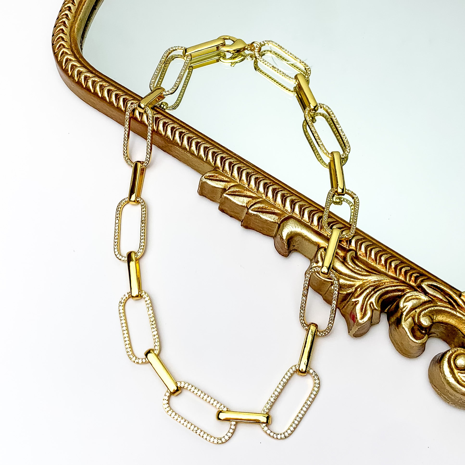 Pictured is a gold chain necklace with alternating chains encrusted with clear crystals. This necklace is pictured partially laying on a gold mirror on a white background.  