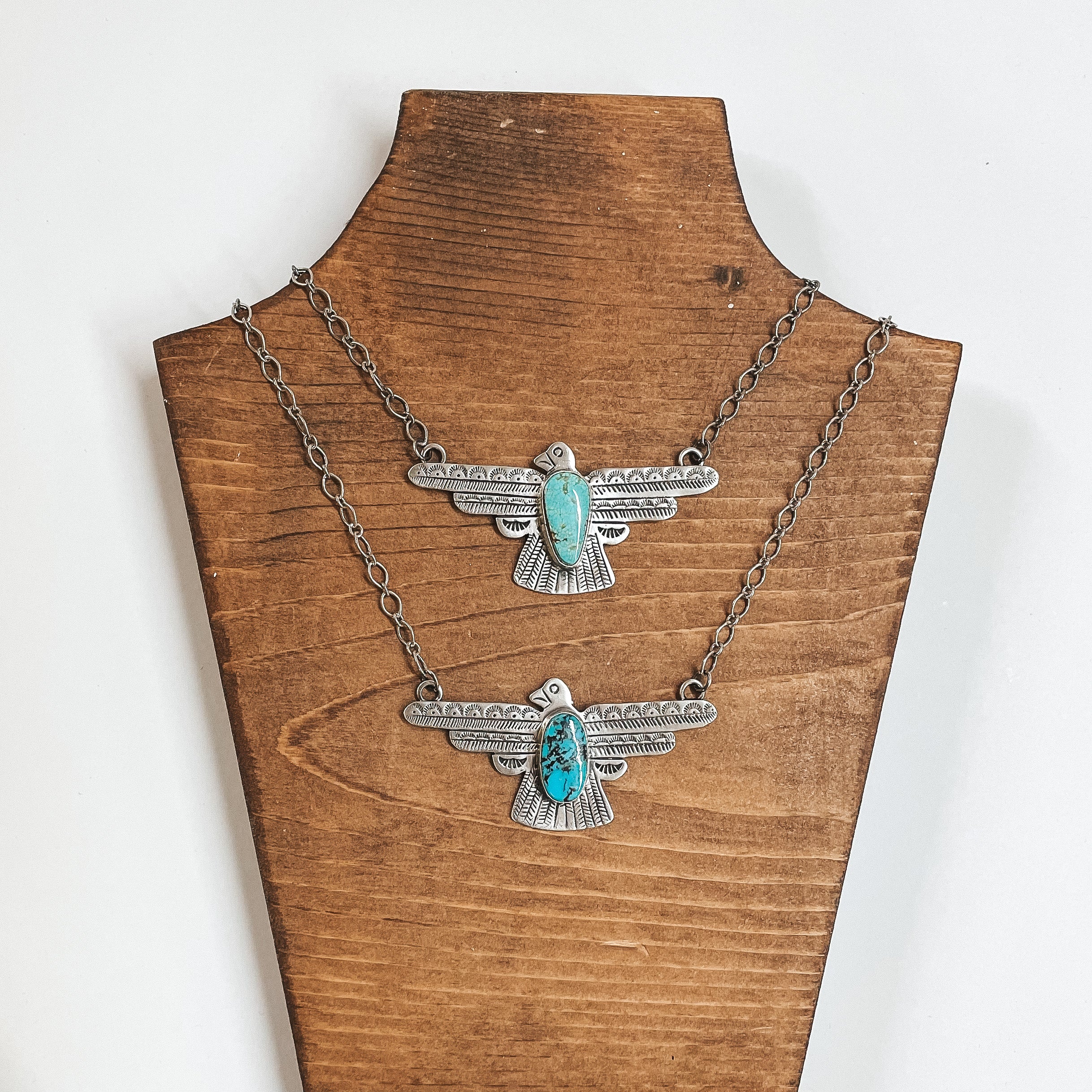 Two silver chain necklaces and they both have thunderbird pendants. Each pendant has a unique shape, size, and shade turquoise stone. These necklaces are pictured on a white background with silver beads under the necklaces.