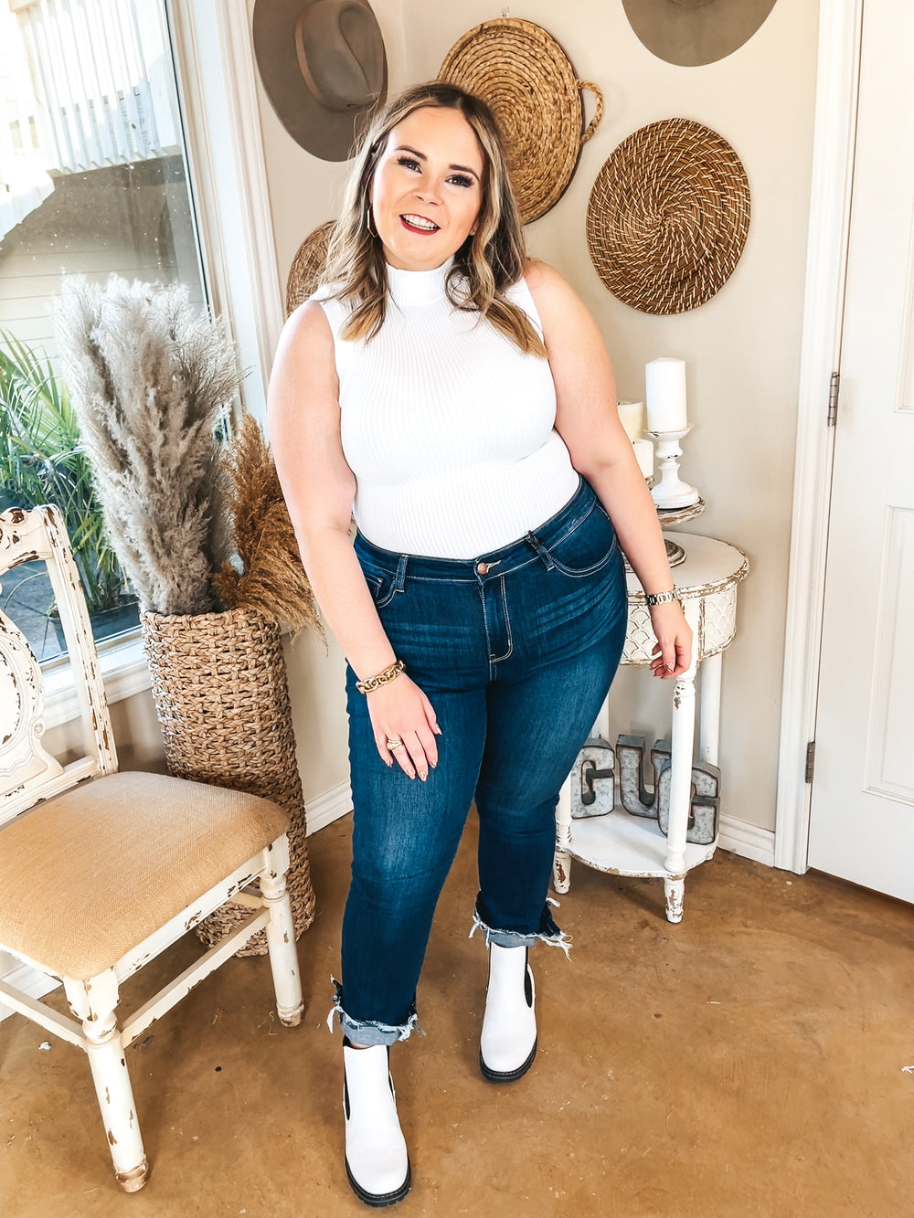 Model is wearing a white, ribbed, turtleneck, tank top bodysuit with dark wash skinny jeans. Jeans are rolled up and have a raw hem. She is also wearing white booties, gold bracelets and rings.