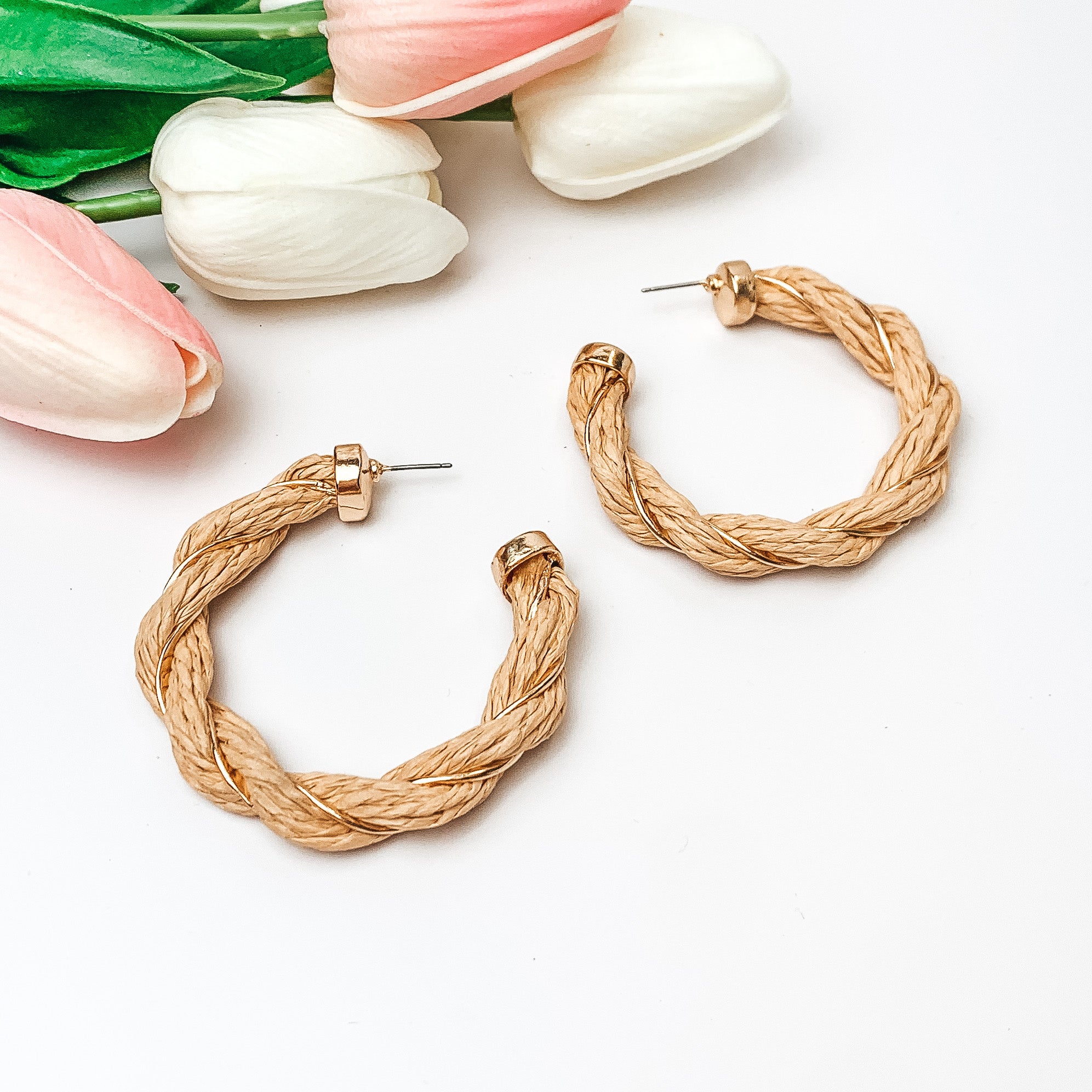 Pictured are beige raffia twisted hoop earrings with gold detailing.  They are pictured with pink and white tulips on a white background.