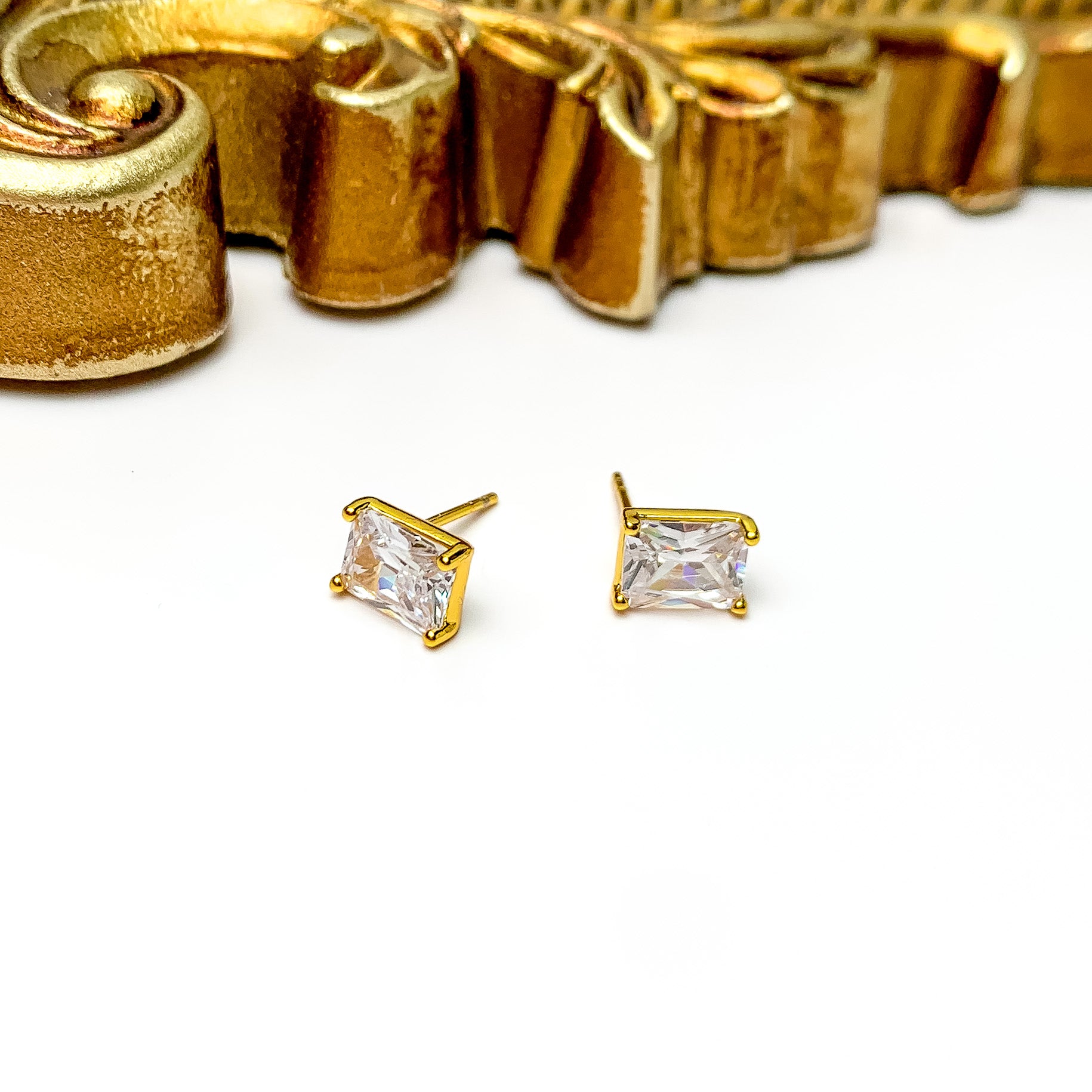 A pair of rectangle, clear crystal stud earrings with a gold setting. These earrings are pictured on a white background with a gold material at the top of the picture. 