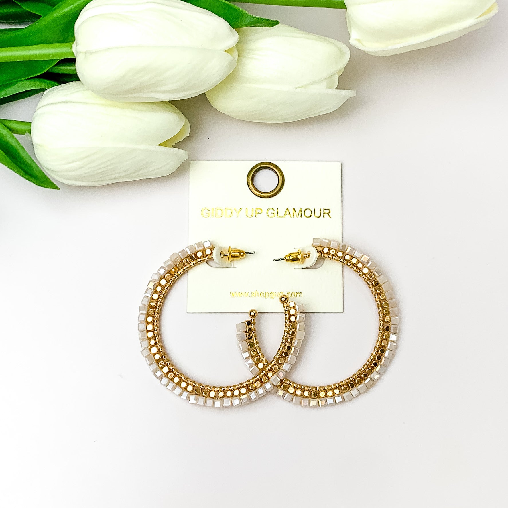 Pictured are circle gold toned hoop earrings with gold beads around it and a palr pink crystal outline. They are pictured with white flowers on a white background.