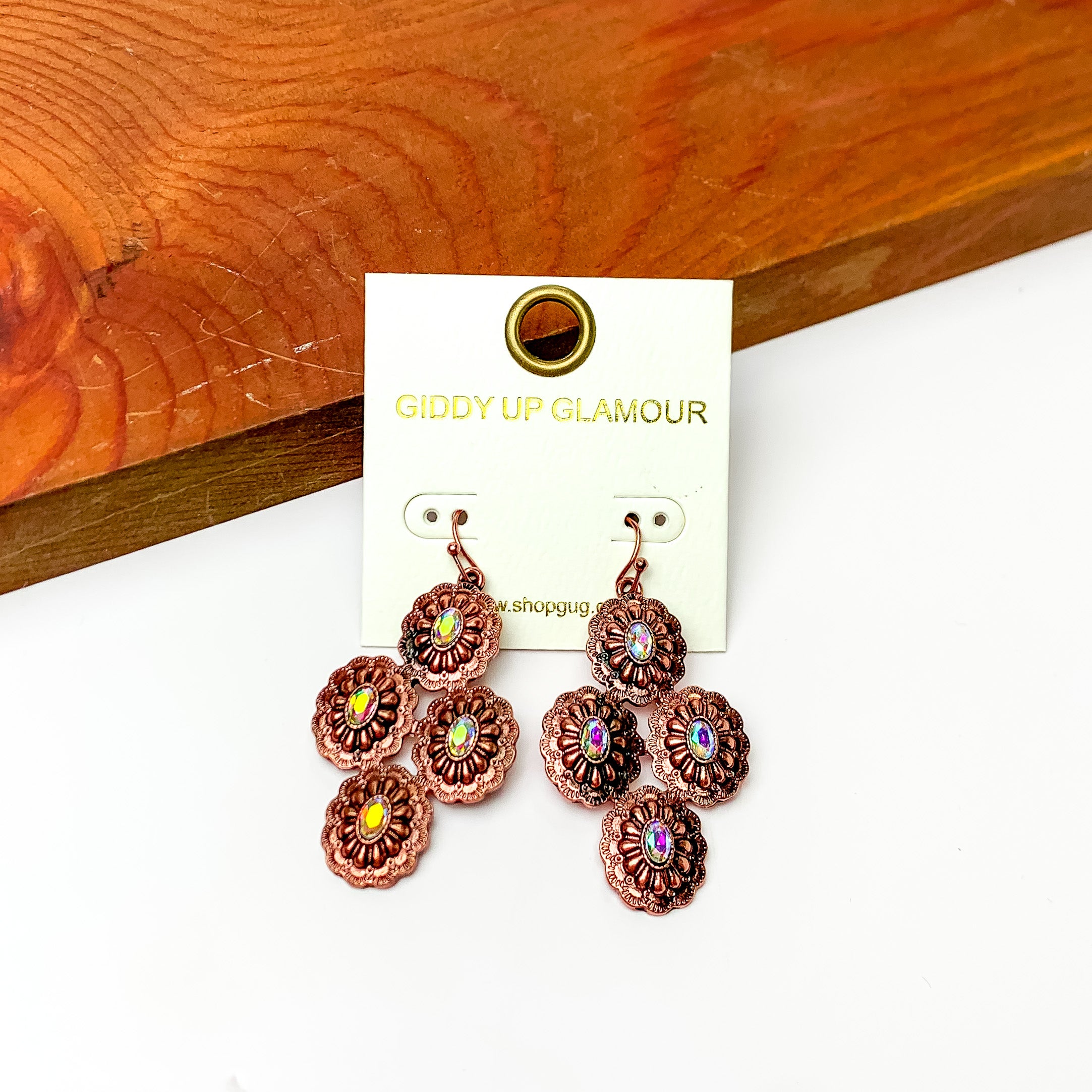 Copper tone flower design drop earrings with ab crystals. Pictured on a white background with a wood piece at the top.