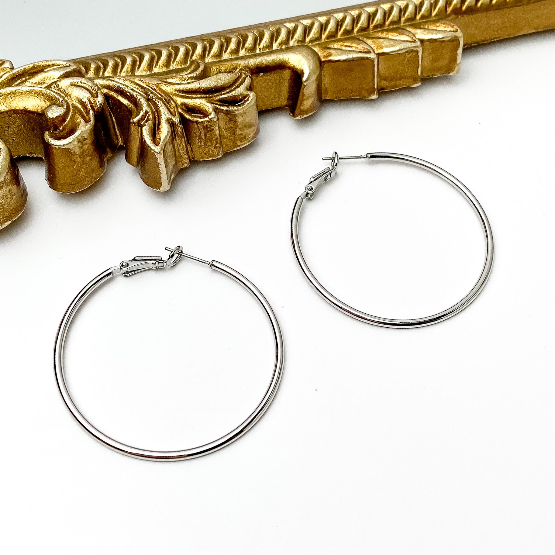Pictured are a pair of silver hoop earrings. These earrings are pictured in front of a gold mirror on a white background. 