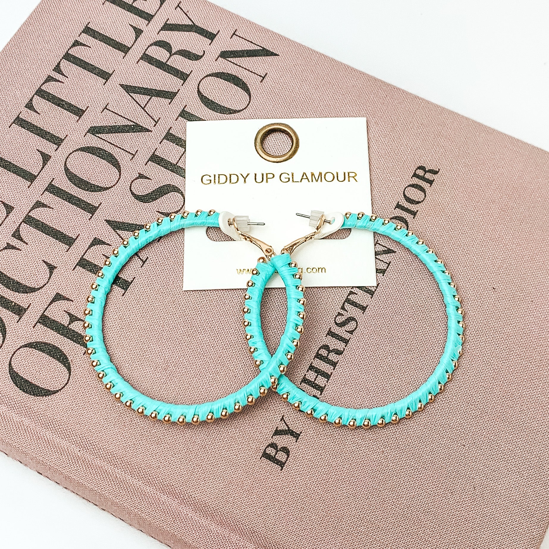 Pictured are circle light blue hoop earrings with gold beads around it. They are pictured with a pink fashion journal on a white background.