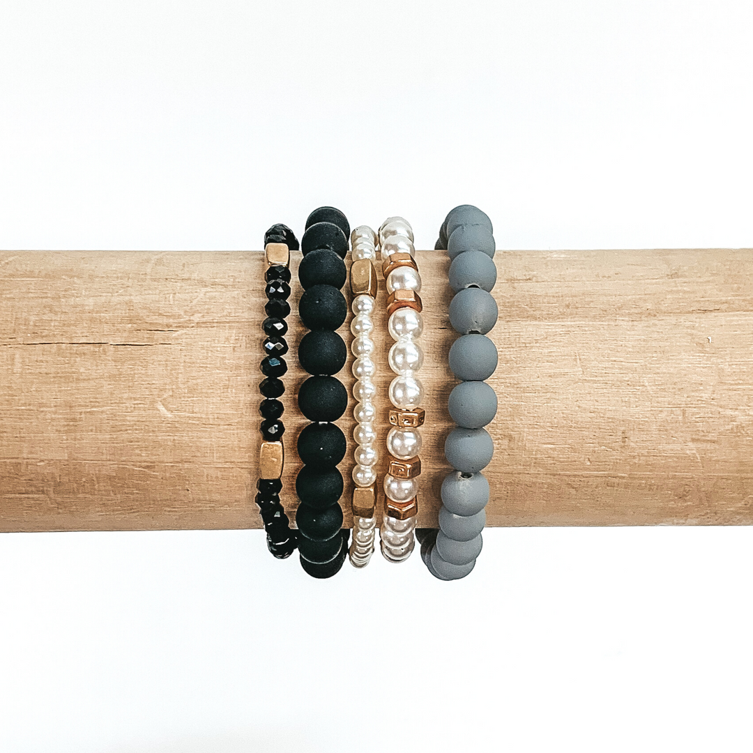 Beaded bracelet set with five different bracelets. Two are matte beaded bracelets with one in black and one in grey. Two bracelets include white pearls with gold or rose gold spacers throughout. The last bracelet has black crystal beads with gold spacers. these bracelets are pictured on a wood bracelet holder on a white background.