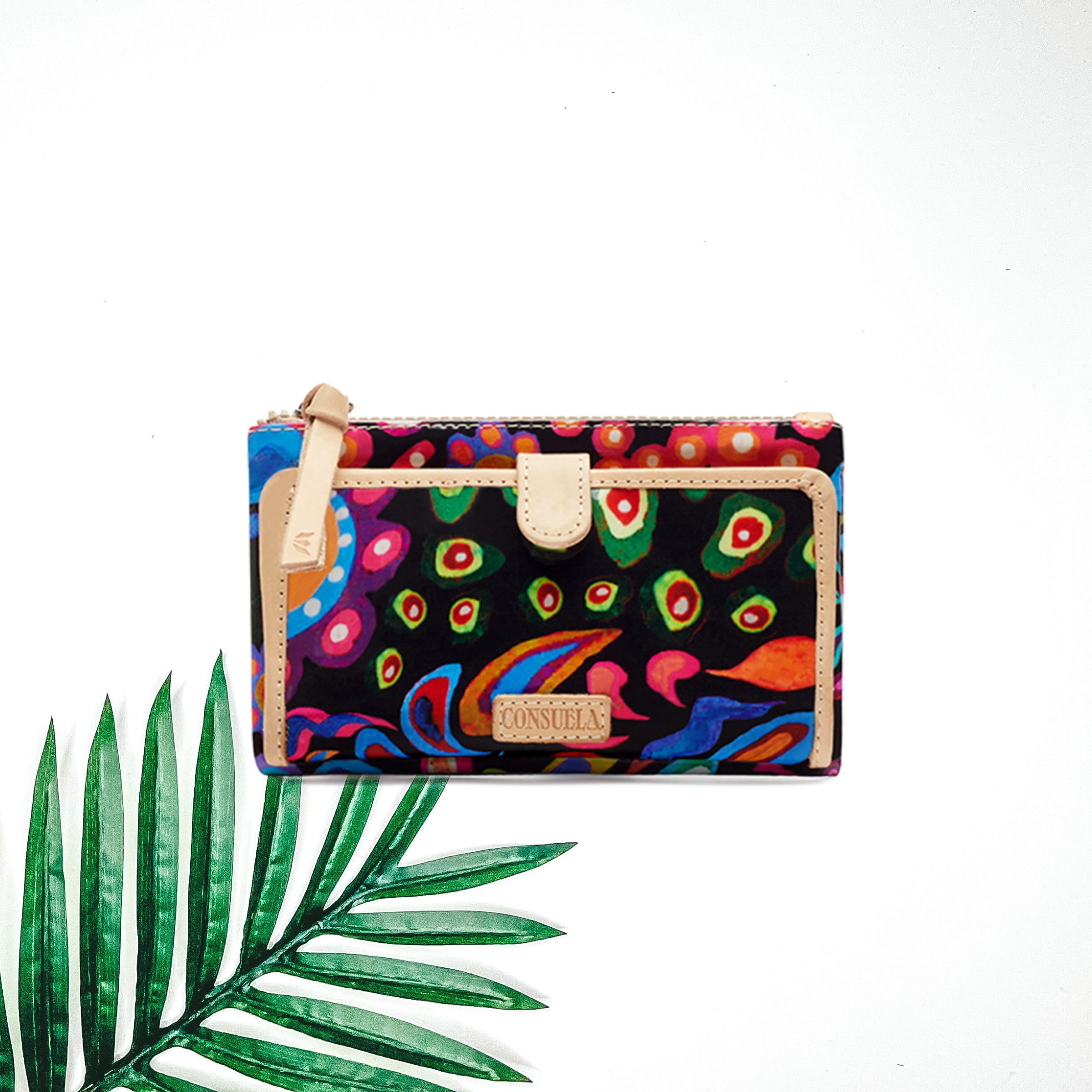 Centered in the picture is a slim wallet with various colored designs. To the left of the wallet is a palm leaf, all on a white background. 