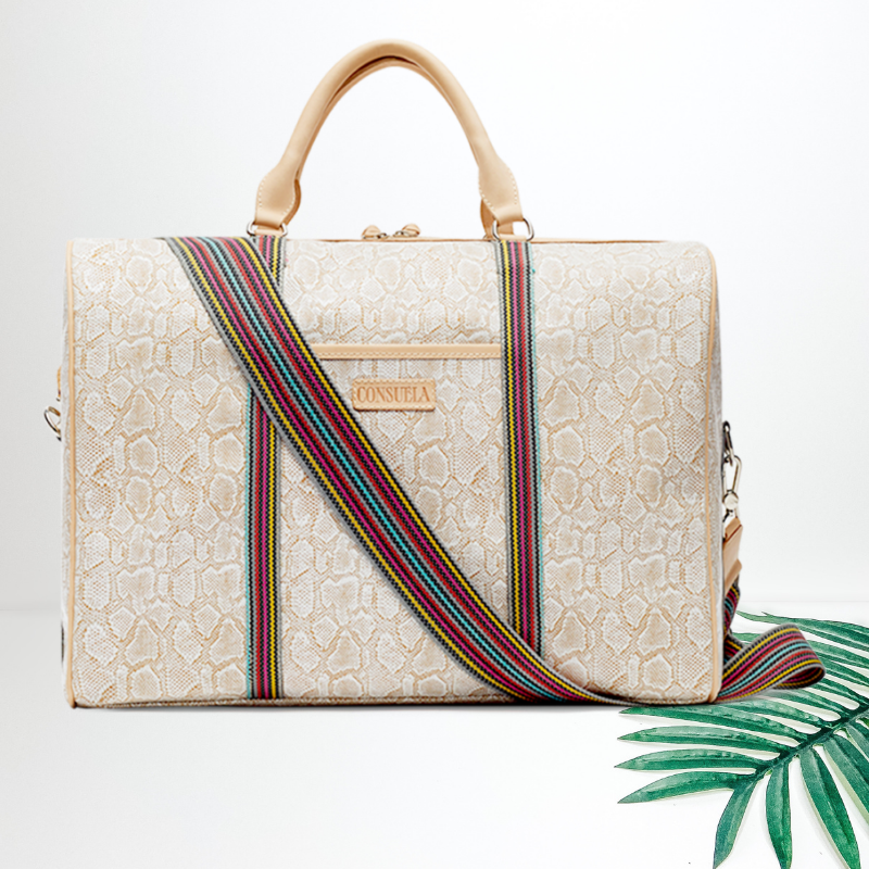 Centered in the picture is a large duffle bag in a cream and white snakeskin. To the right of the bag is a palm leaf, all on a white background. 
