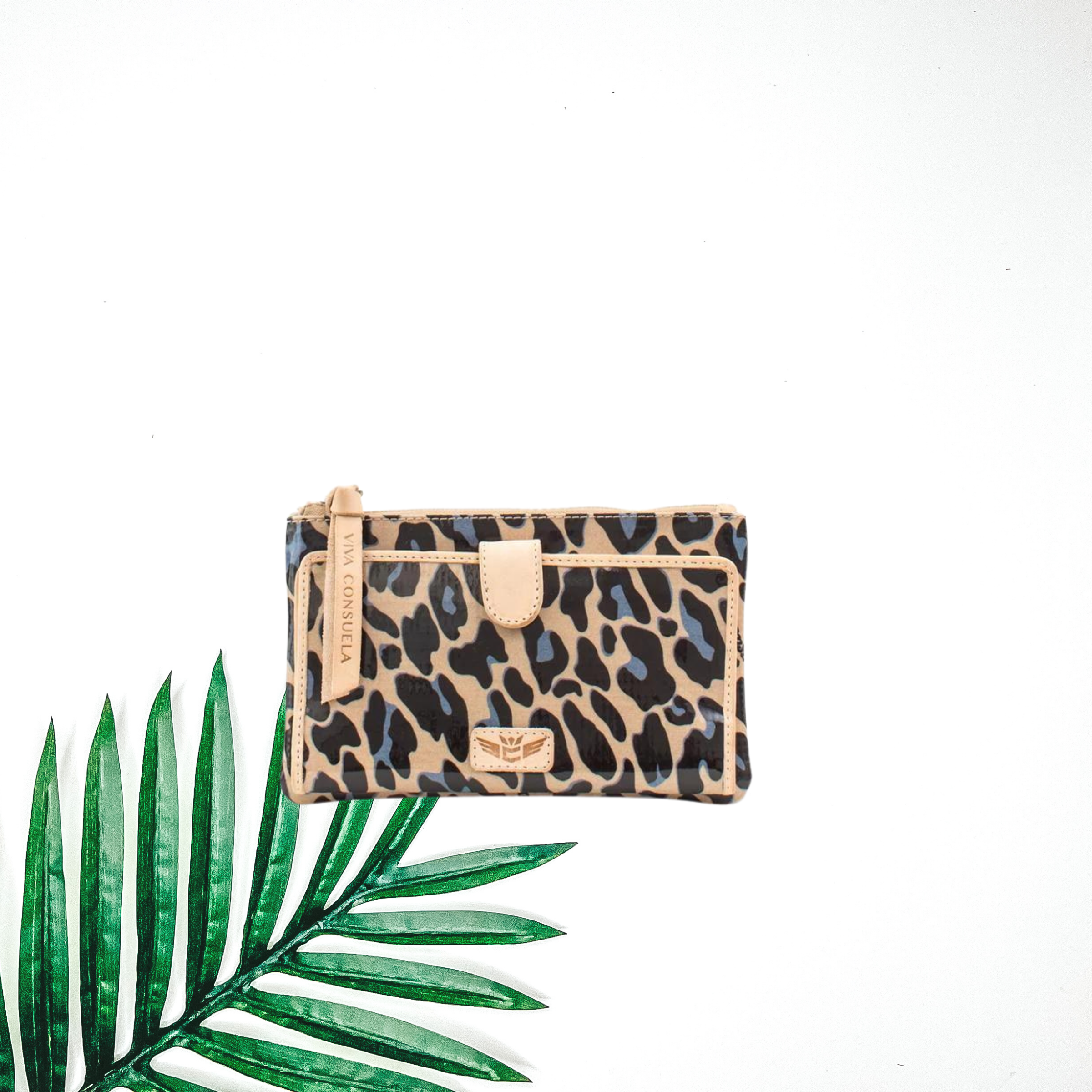 Centered is a cheetah print slim wallet with blue accents. To the right of the wallet is a palm leaf, all on a white background. 