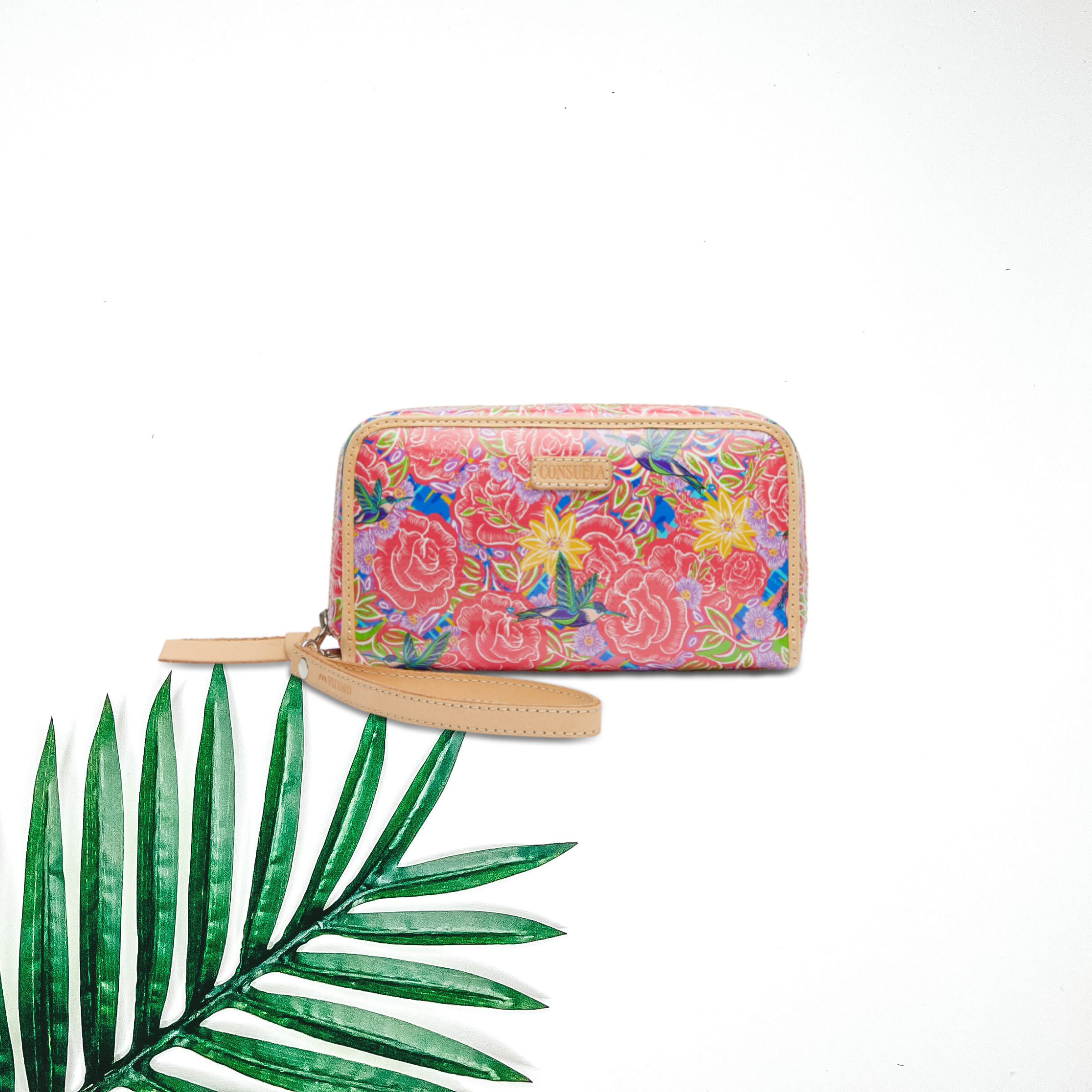 Centered in the picture is a multi colored floral wristlet wallet. A palm leaf is on the bottom right corner. Background is white. 