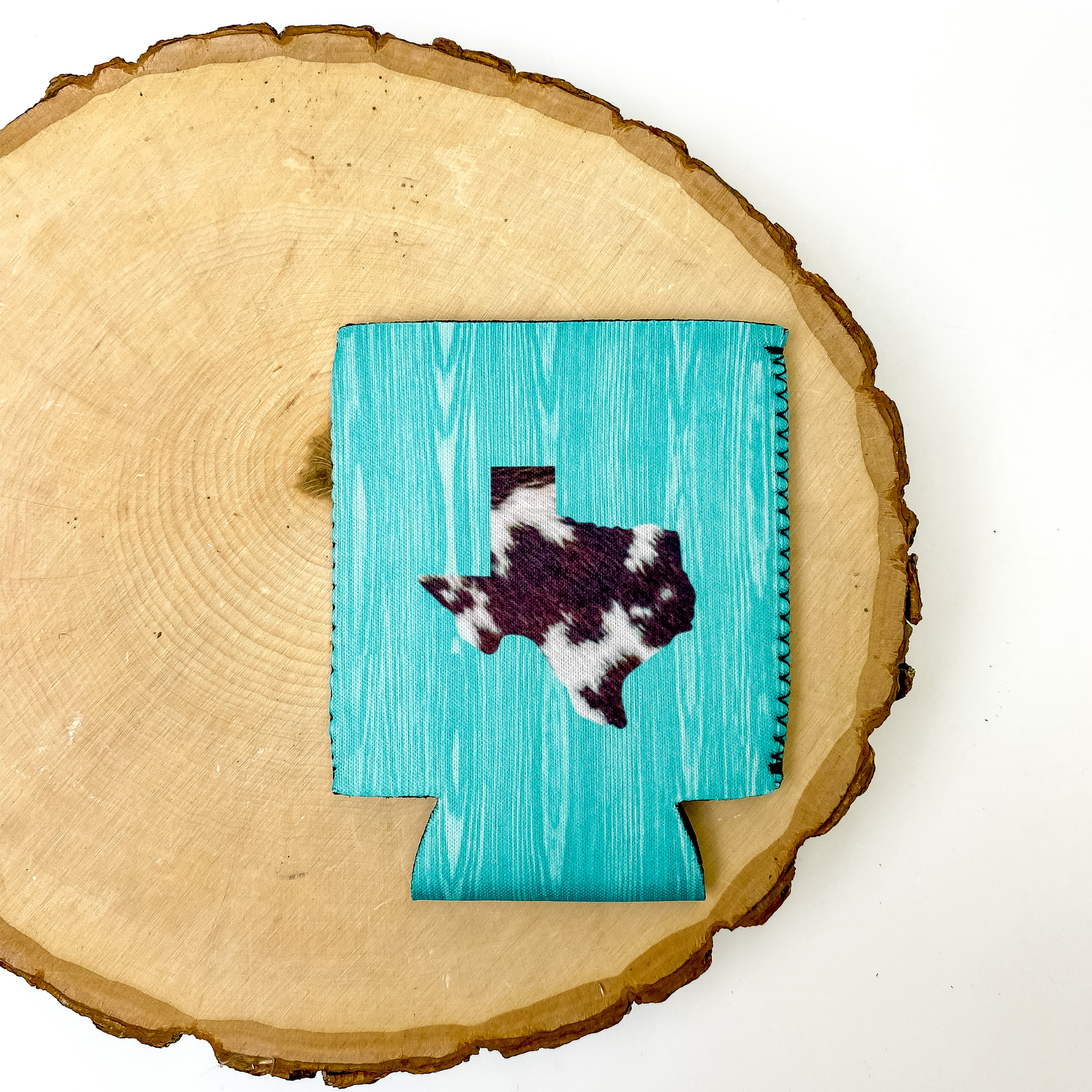 Turquoise wooden print on the background and a cow print texas in the middle of koozie. This koozie is pictured on a piece of wood on a white background.