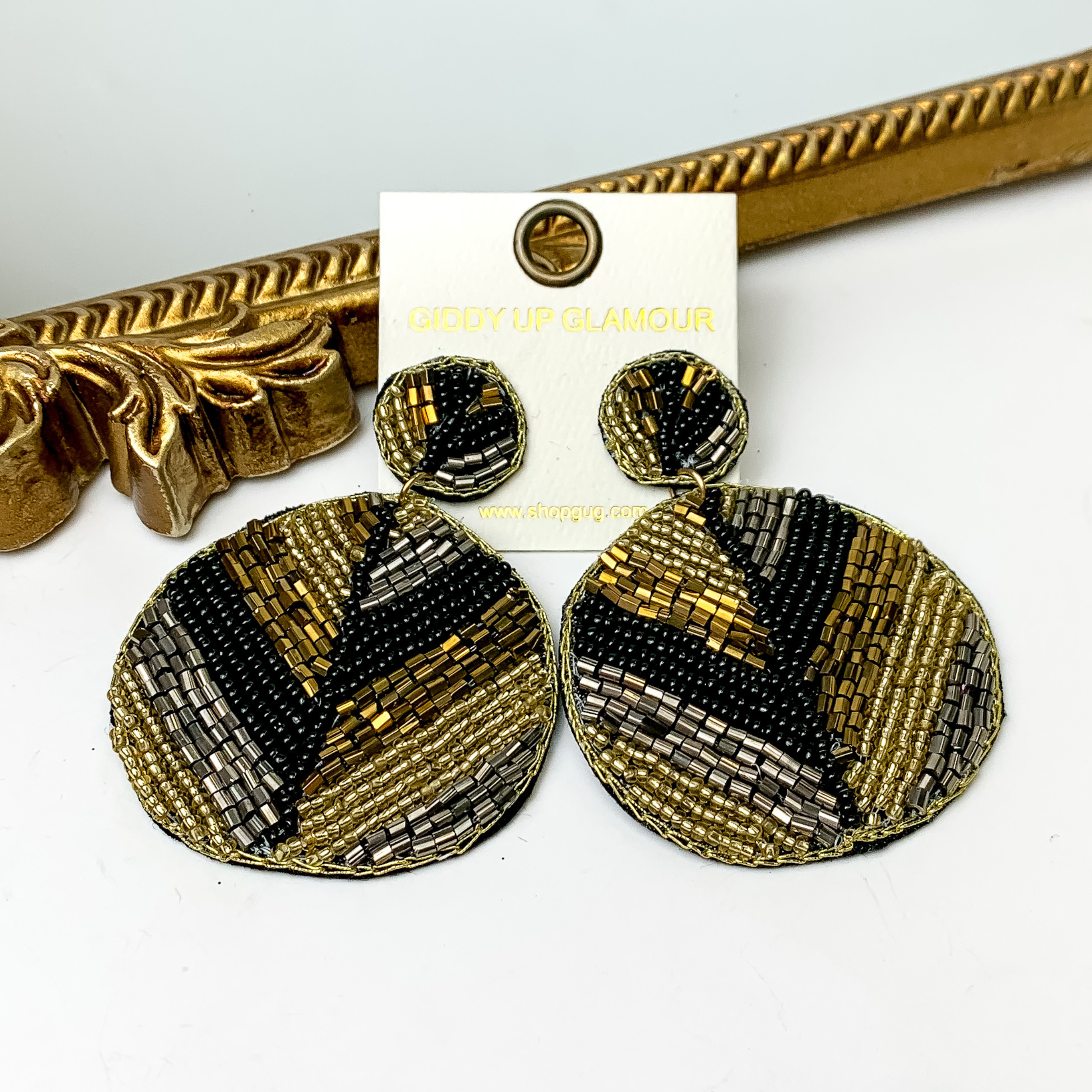 Pictured is a pair of beaded circle drop earrings in a V-shaped stripe pattern. These earrings include black, gold, bronze, and silver beads. These earrings are pictured in a white background with a gold mirror.