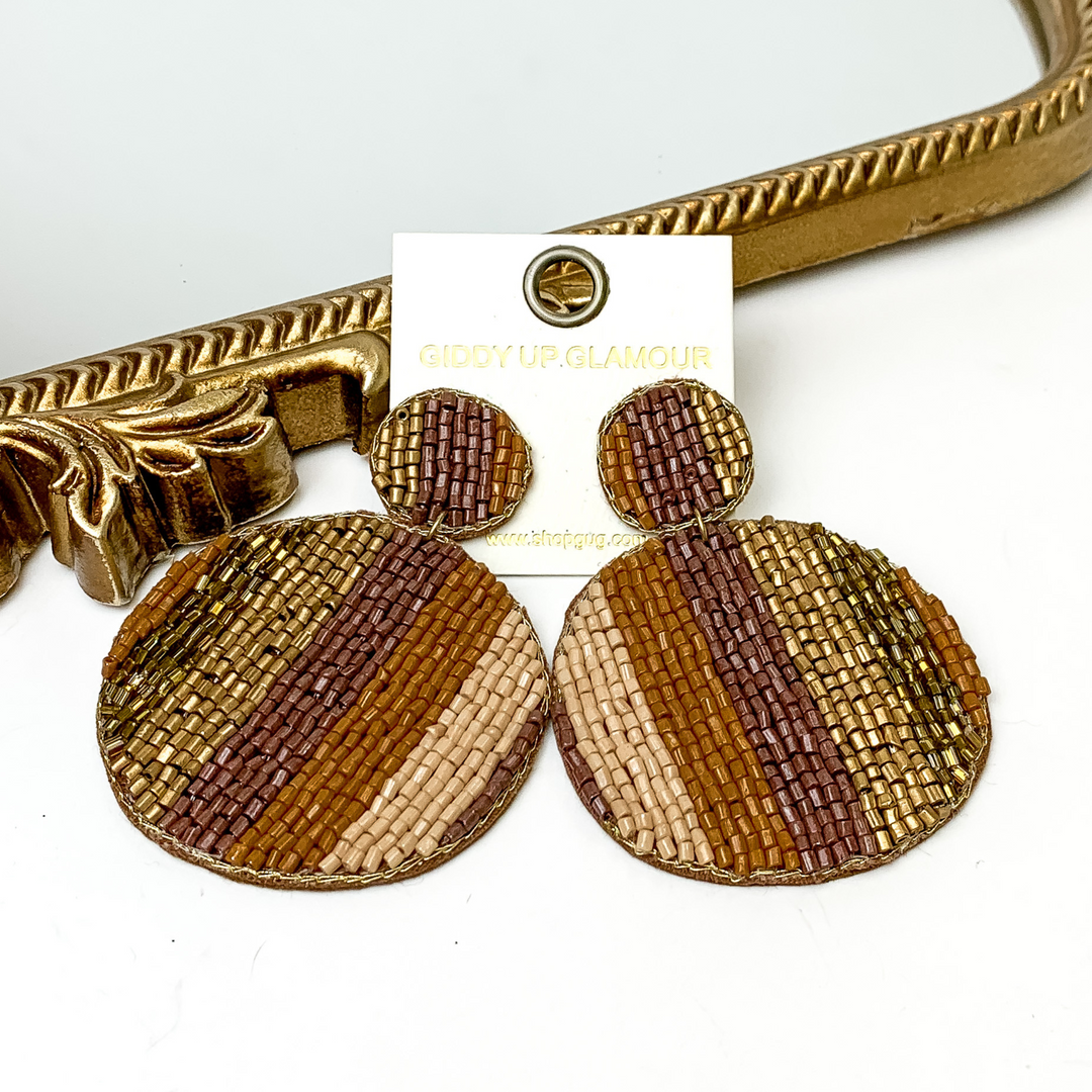 Pictured is a pair of beaded circle drop earrings in a stripe pattern. These earrings include multi colors of brown. These earrings are pictured in a white background with a gold mirror.