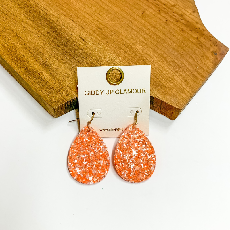 Pictured is orange glitter teardrop earrings. These earrings are pictured on a white background with a wooden board.