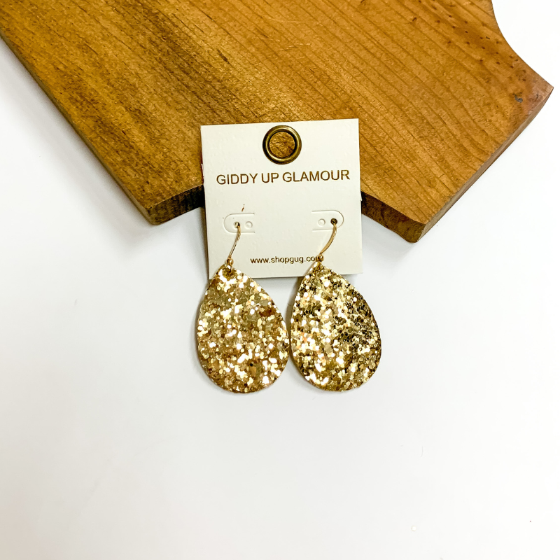 Pictured is gold glitter teardrop earrings. These earrings are pictured on a white background with a wooden board.