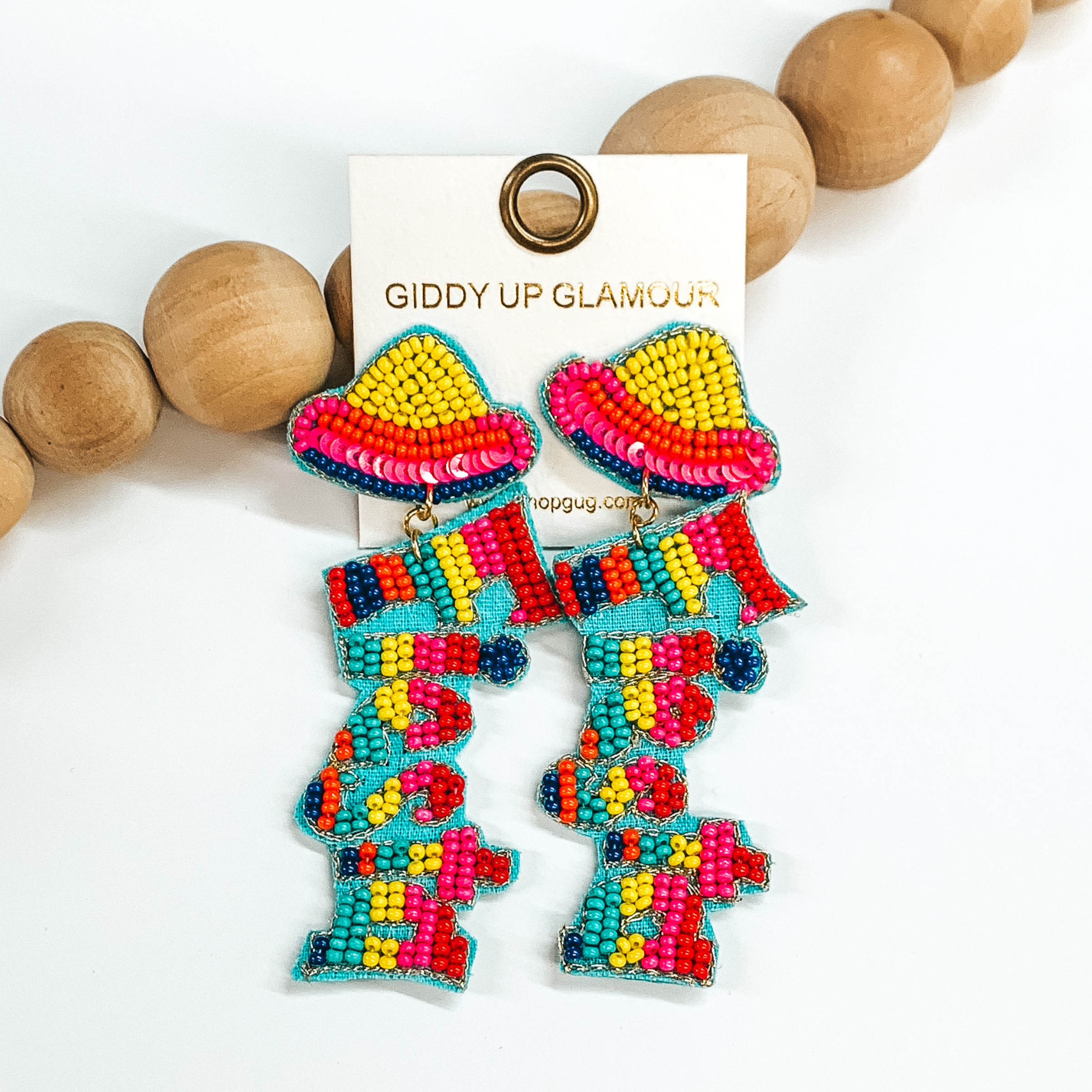Sombrero fiesta seed beaded drop earrings in  multicolor. The sombrero has yellow, orange, dark  blue, and pink sequins. The fiesta has red, pink,  yellow, turquoise, orange, dark blue, and  red beads. Outlined in turquoise with gold thread. Taken in a white background with brown beads as decor.