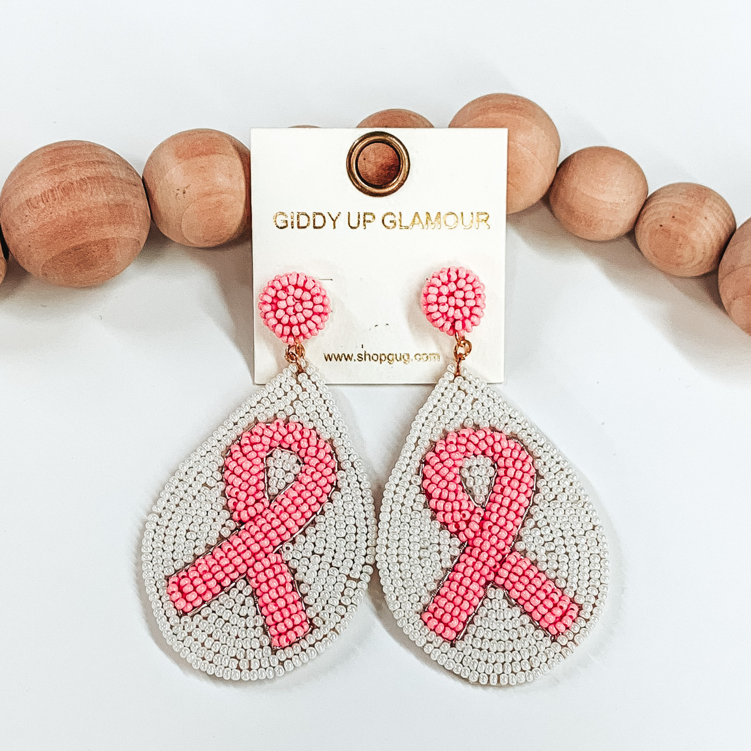 Light pink beaded circle studs with hanging beaded teardrop pendant. The pendant has a  white beaded background with a center beaded  light pink ribbon These earrings are pictured on a  white background with brown beads as decor. 