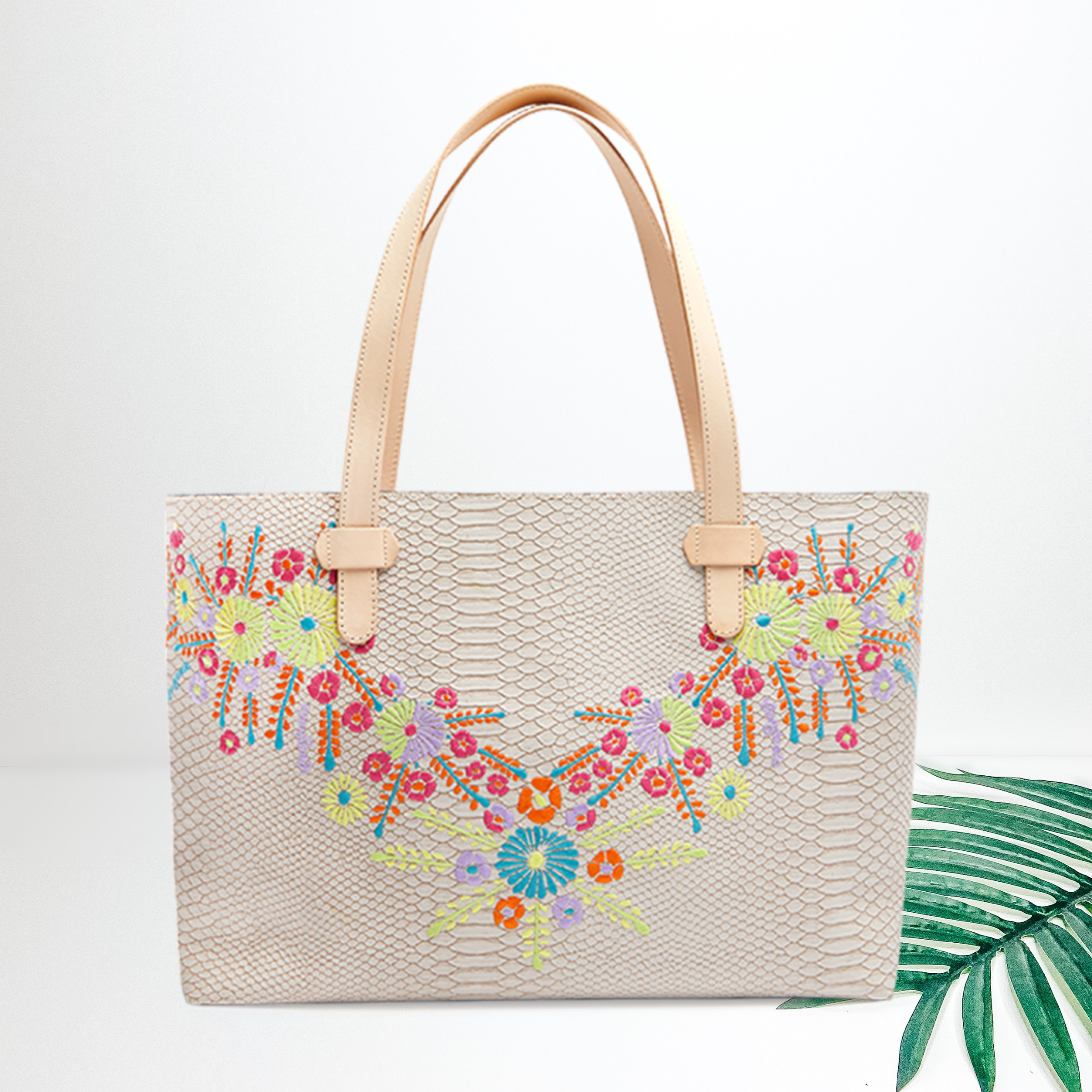Centered in the picture is a large tote that has yellow, orange, blue, purple and pink embroidery along the sides. To the right of the tote is a palm leaf on a white background. 