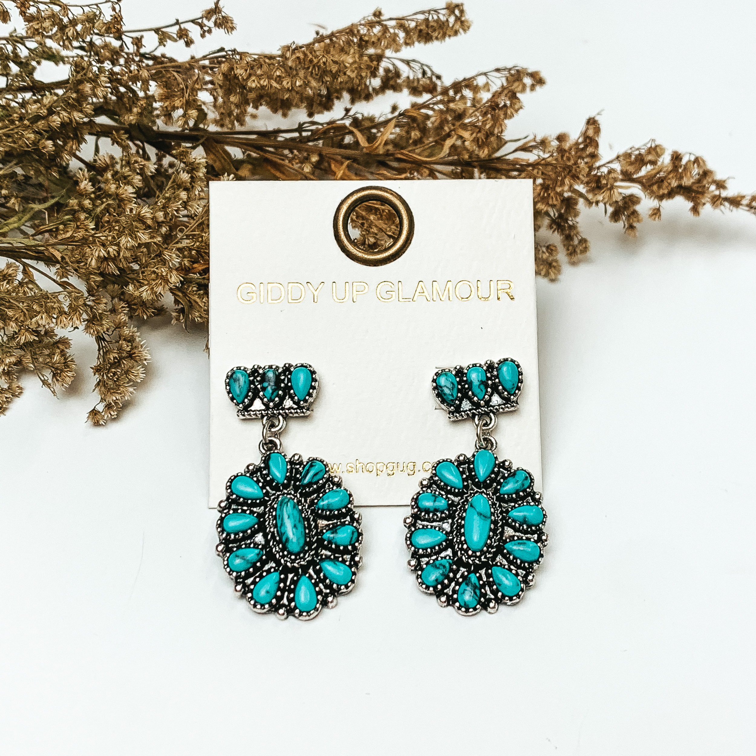 Silver tone cluster post earrings with oval cluster drop with turquoise stones. These earrings are pictured on a white background with greenery behind the earrings.