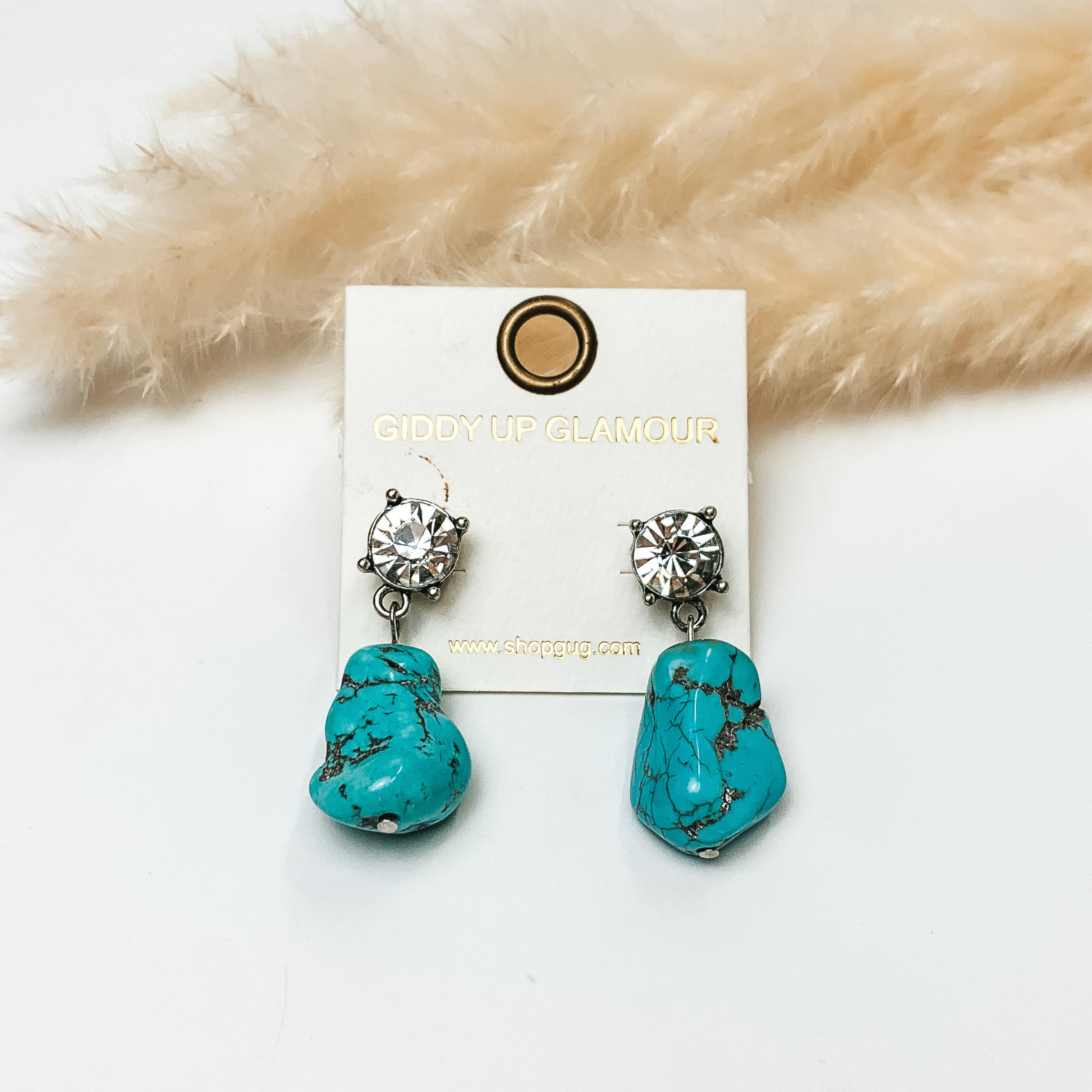 Clear crystal post back earrings with large turquoise stones attached. Earrings are in front of greenery with white background. 