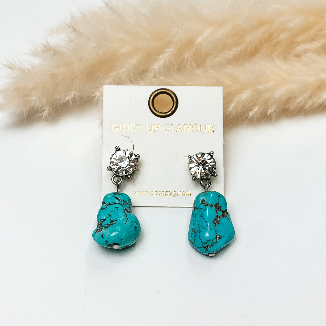 Clear crystal post back earrings with large turquoise stones attached. Earrings are in front of greenery with white background. 