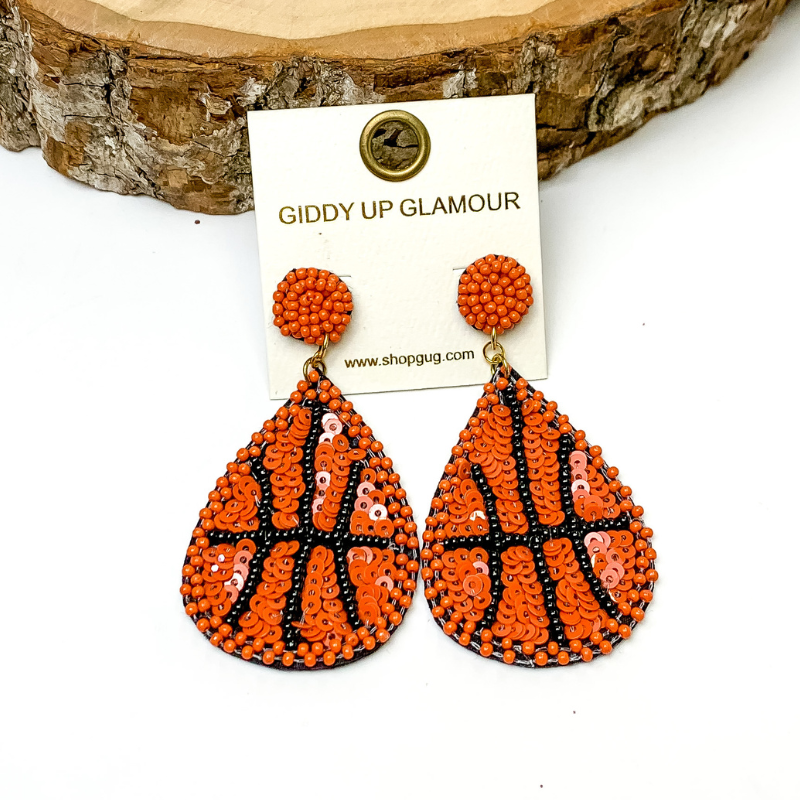 Pictured is teardrop beaded sequin basketball earrings in orange. They are propped up on a piece of circular wood on a white background.