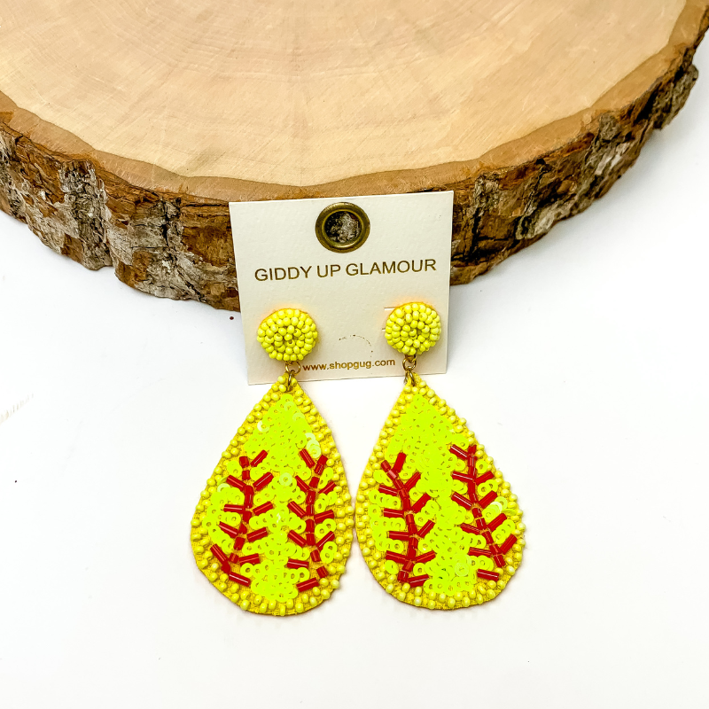 Pictured are sequin teardrop softball earrings with circle beaded post and red beaded detailing and neon yellow sequins. They are propped up on a circle piece of wood on a white background.