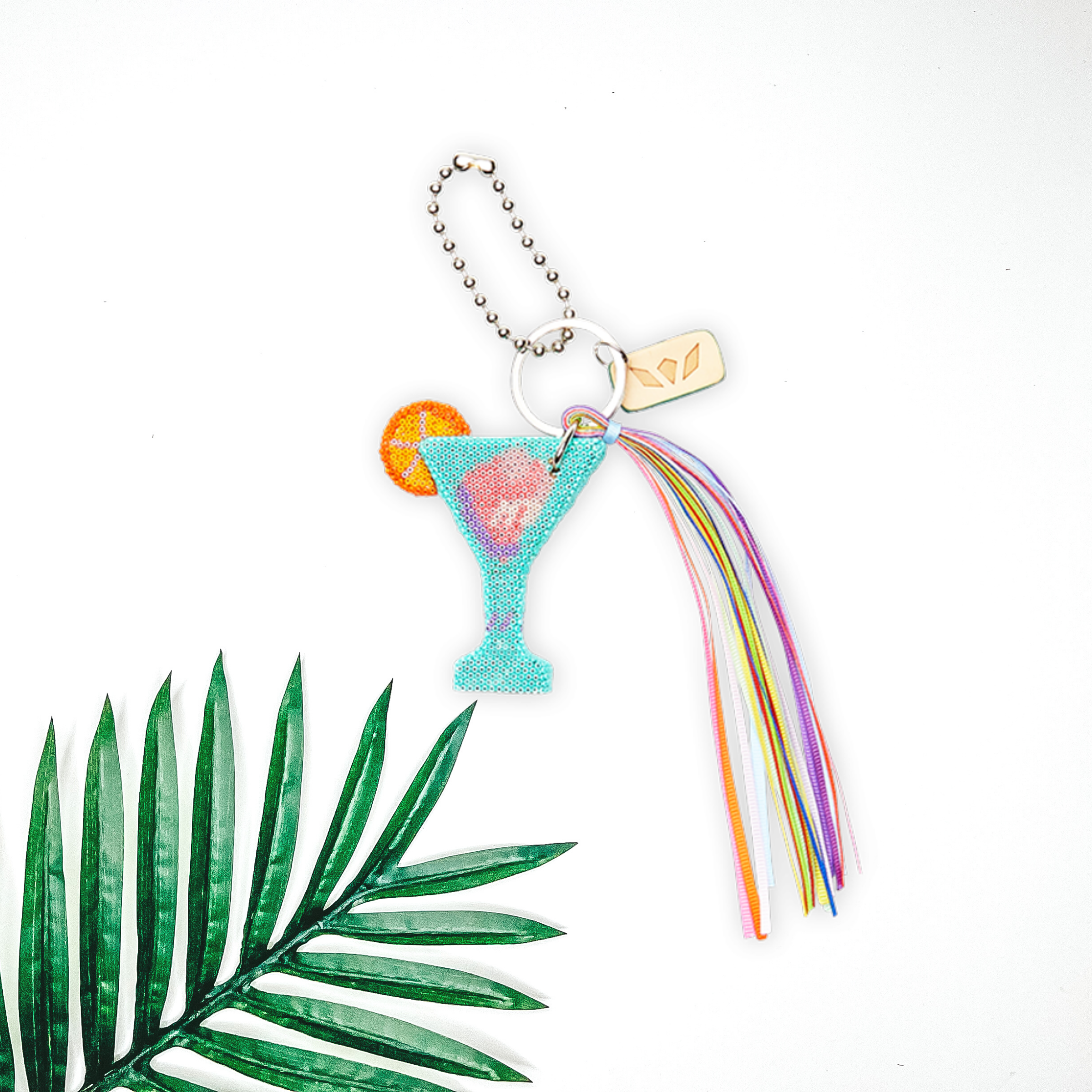 Centered in the middle of the picture is a beaded butterfly charm with multi-colored tassels. To the right of the charm is a palm leaf, all on a white background. 