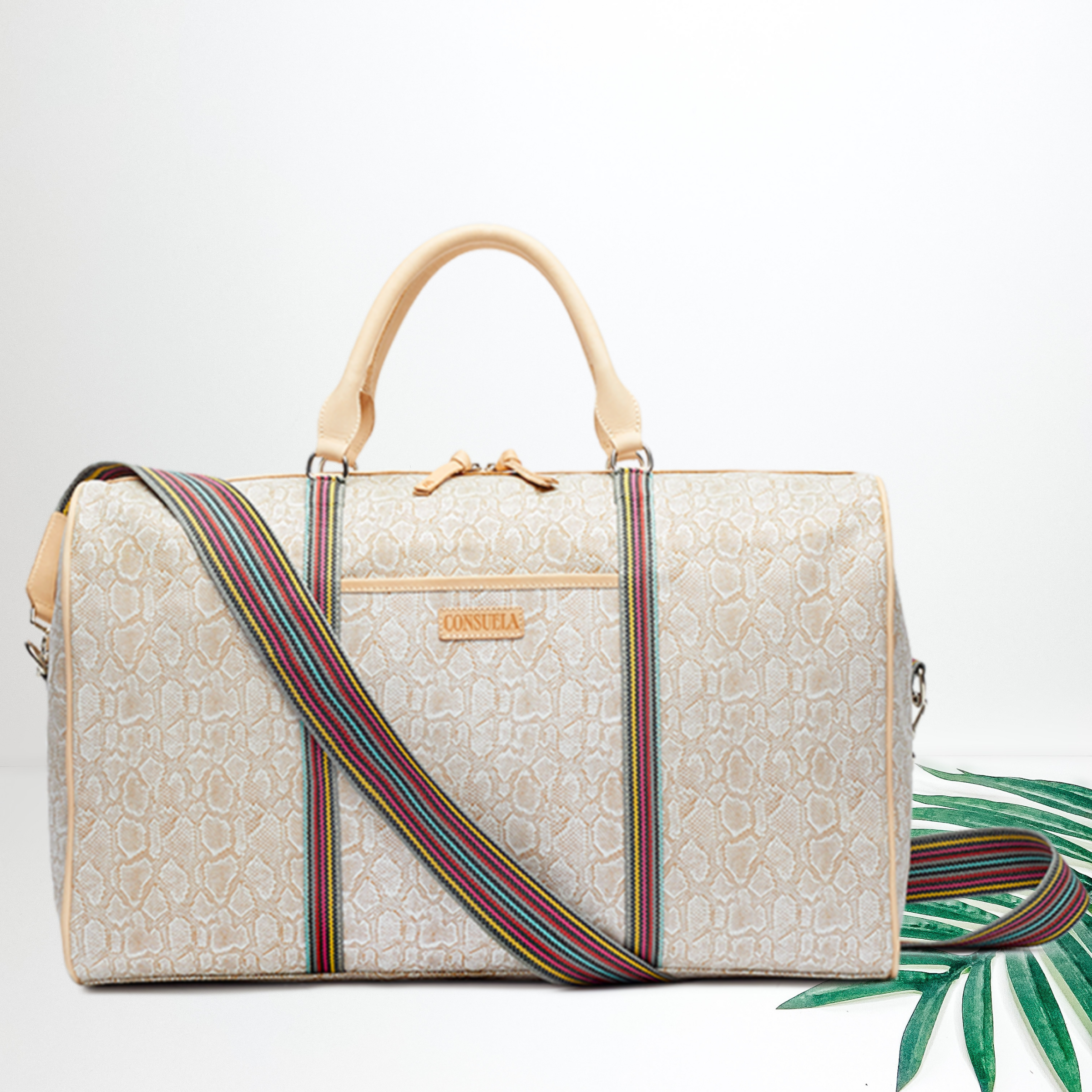Centered in the picture is a tote bag in white snakeskin. To the right of the tote is a palm leaf, all on a white background.