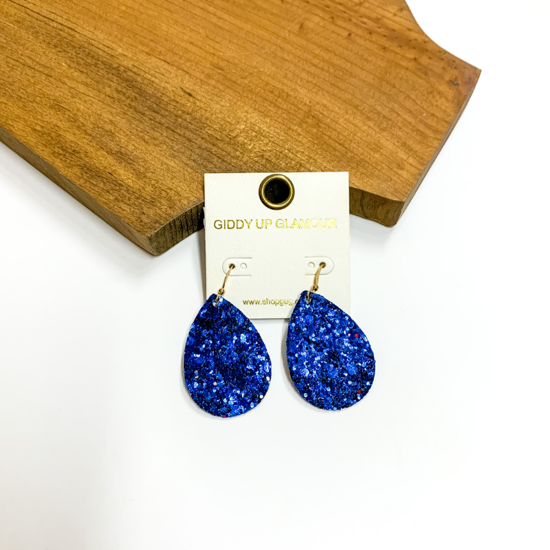 Pictured is blue glitter teardrop earrings. These earrings are pictured on a white background with a wooden board.