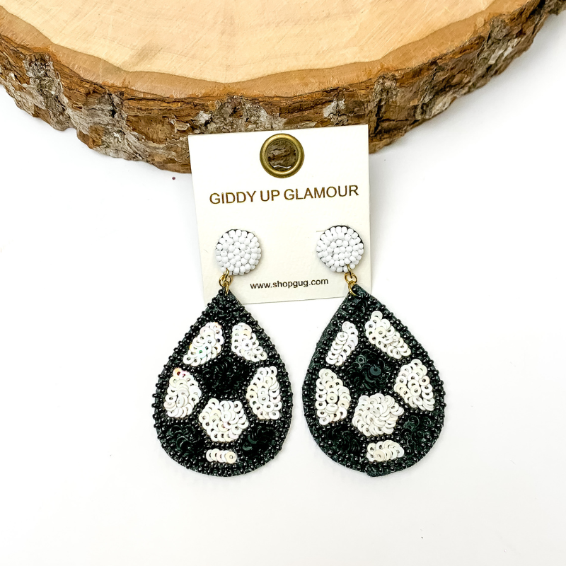 Pictured are teardrop sequin soccerball earrings with beaded detailing. These earrings are propped up on a circle piece of wood on a black background.