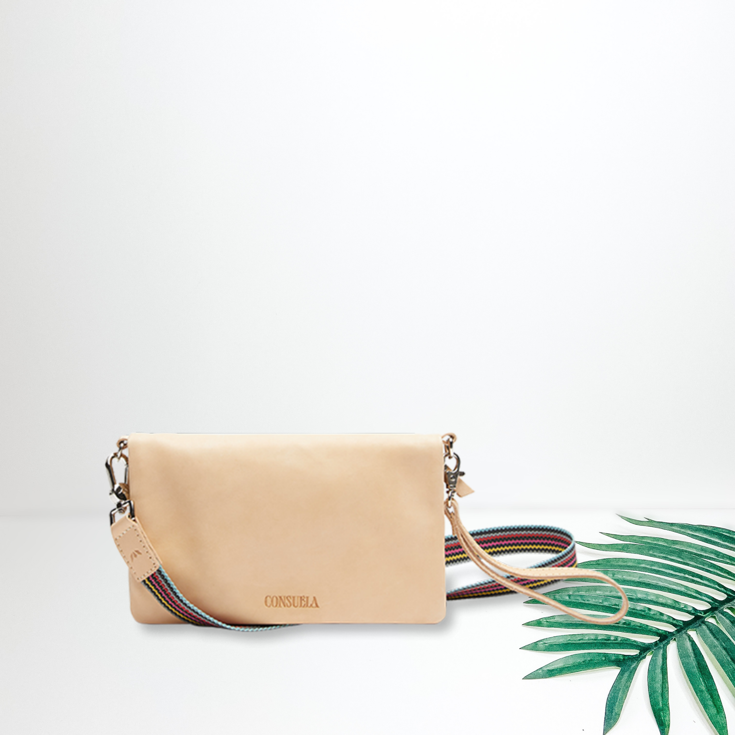 Centered in the picture is a crossbody purse in nude leather. To the right of the bag is a palm leaf, all on a white background.