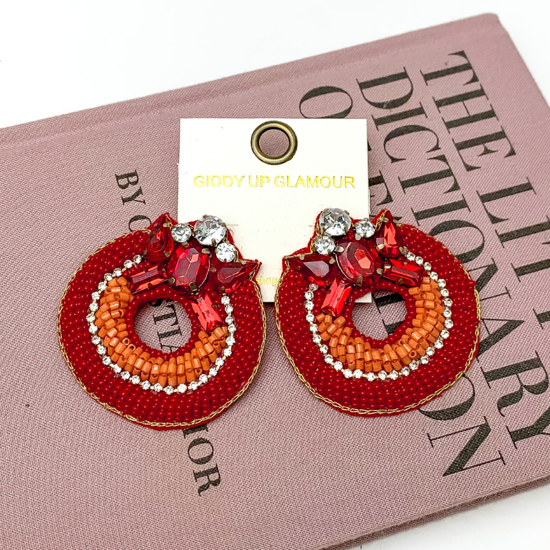 Pictured is hoop beaded red and orange earrings with a jeweled detailing at the top. These earrings are pictured on a white background with a pink fashion dictionary.