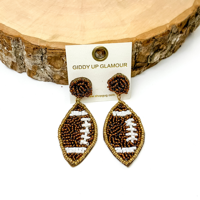 Pictured are beaded footbal earrings in brown with white beaded detailing. These earrings are propped up on a circle piece of wood on a black background