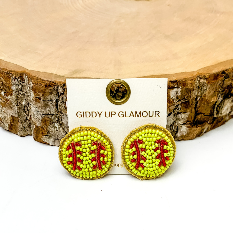 Pictured are beaded yellow softball stud earrings. They are propped up on a piece of circle wood on a white background.