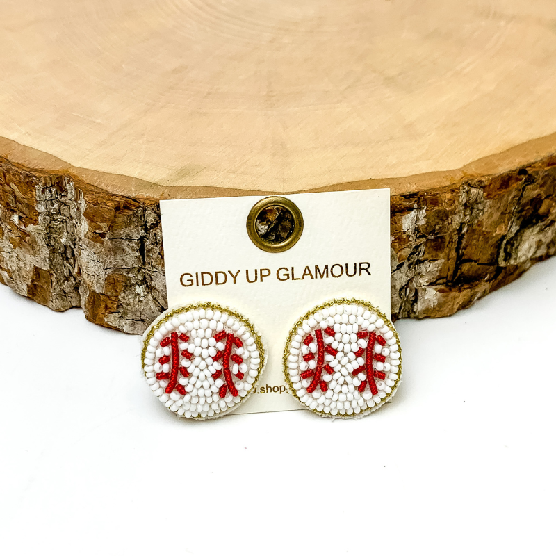 Pictured are beaded white and red baseball stud earrings. They are propped up on a piece of circle wood on a white background.