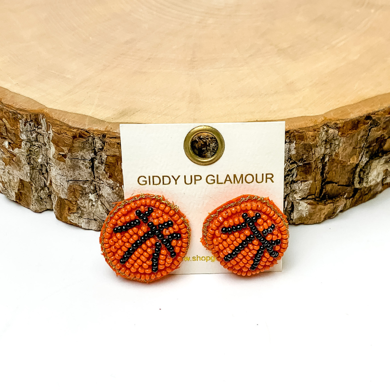 Pictured are beaded orange and black basketballl stud earrings. They are propped up on a piece of circle wood on a white background.