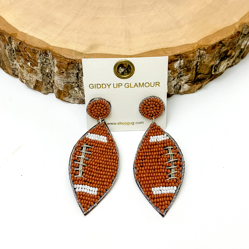Pictured are beaded football earrings in brown with circle beaded post and silver detailing. They are propped up on a circle piece of wood on a white background.