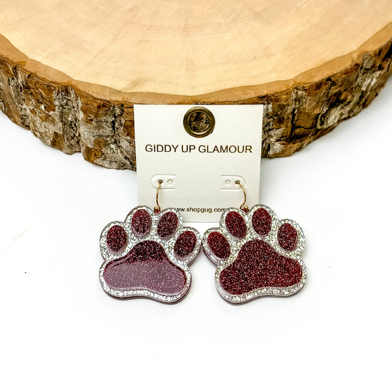 Pictured is maroon paw prints with silver glitter outlining. They are propped up on a wooden circle on a white background. 