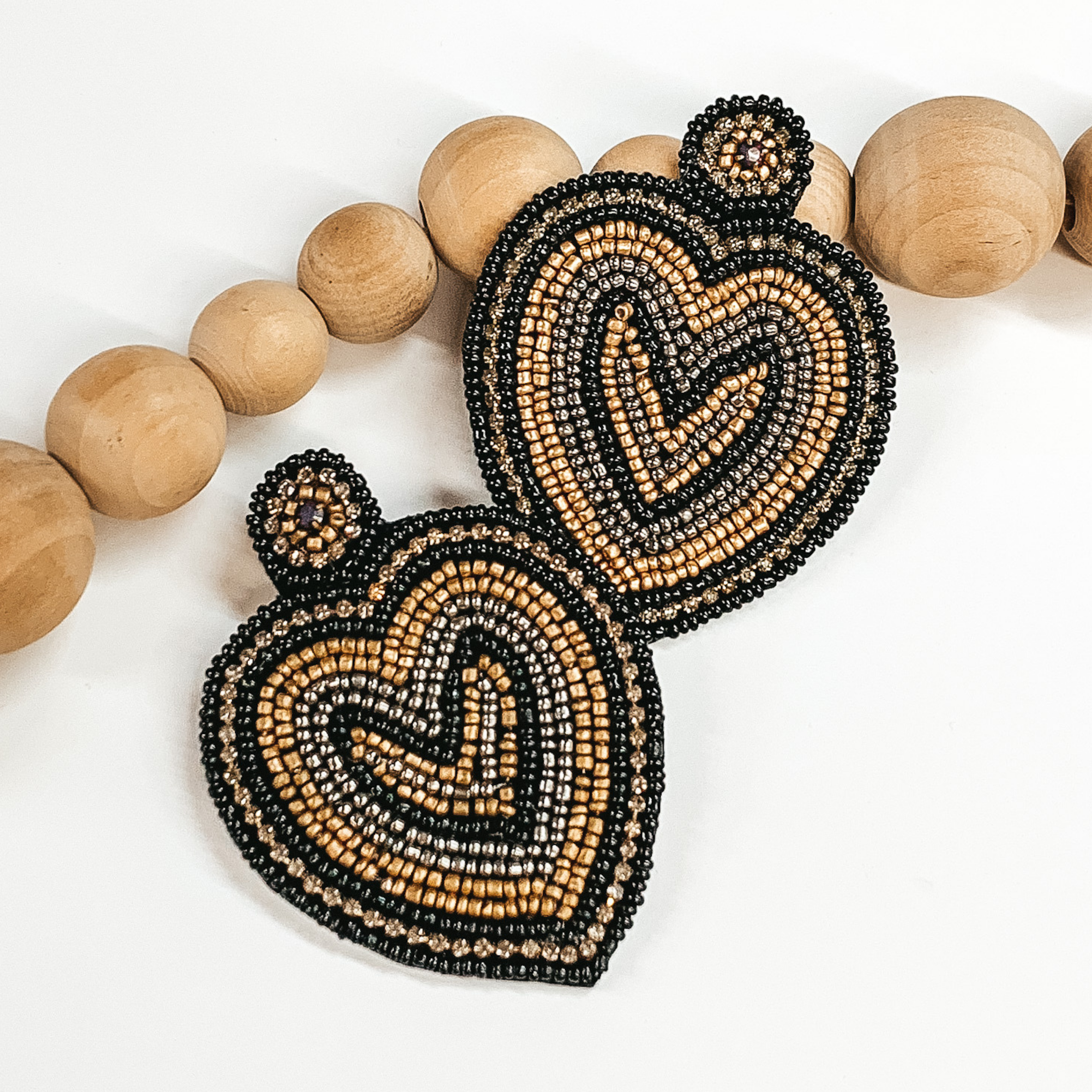 Heart seed beaded earrings in gold, black, silver beads with dark grey crystals in a heart pattern. Taken in a white background with brown beads  as decor.