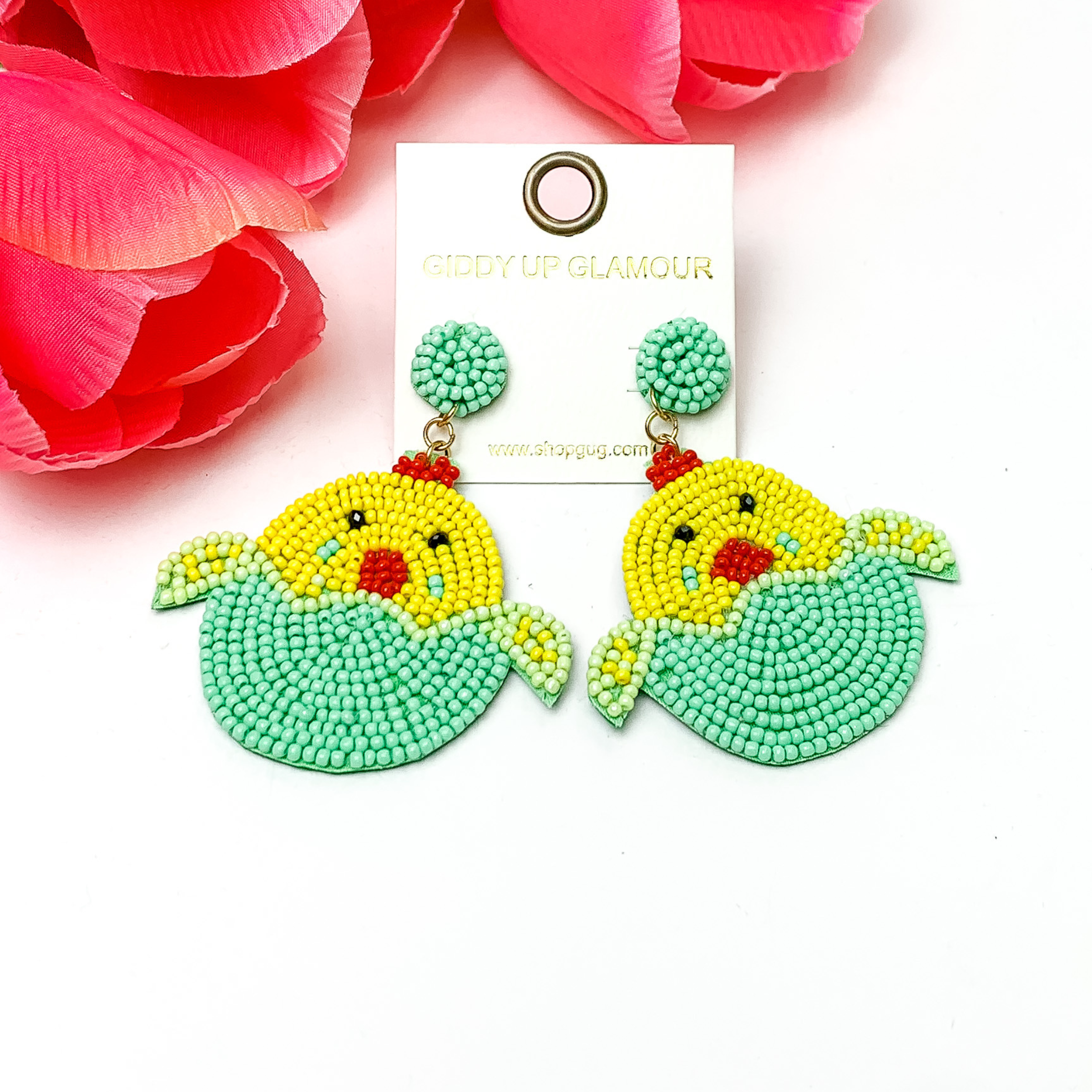 Beaded, mint Easter egg earrings with a yellow beaded chick hatching. Pictured on white background with red-coral colored flowers.
