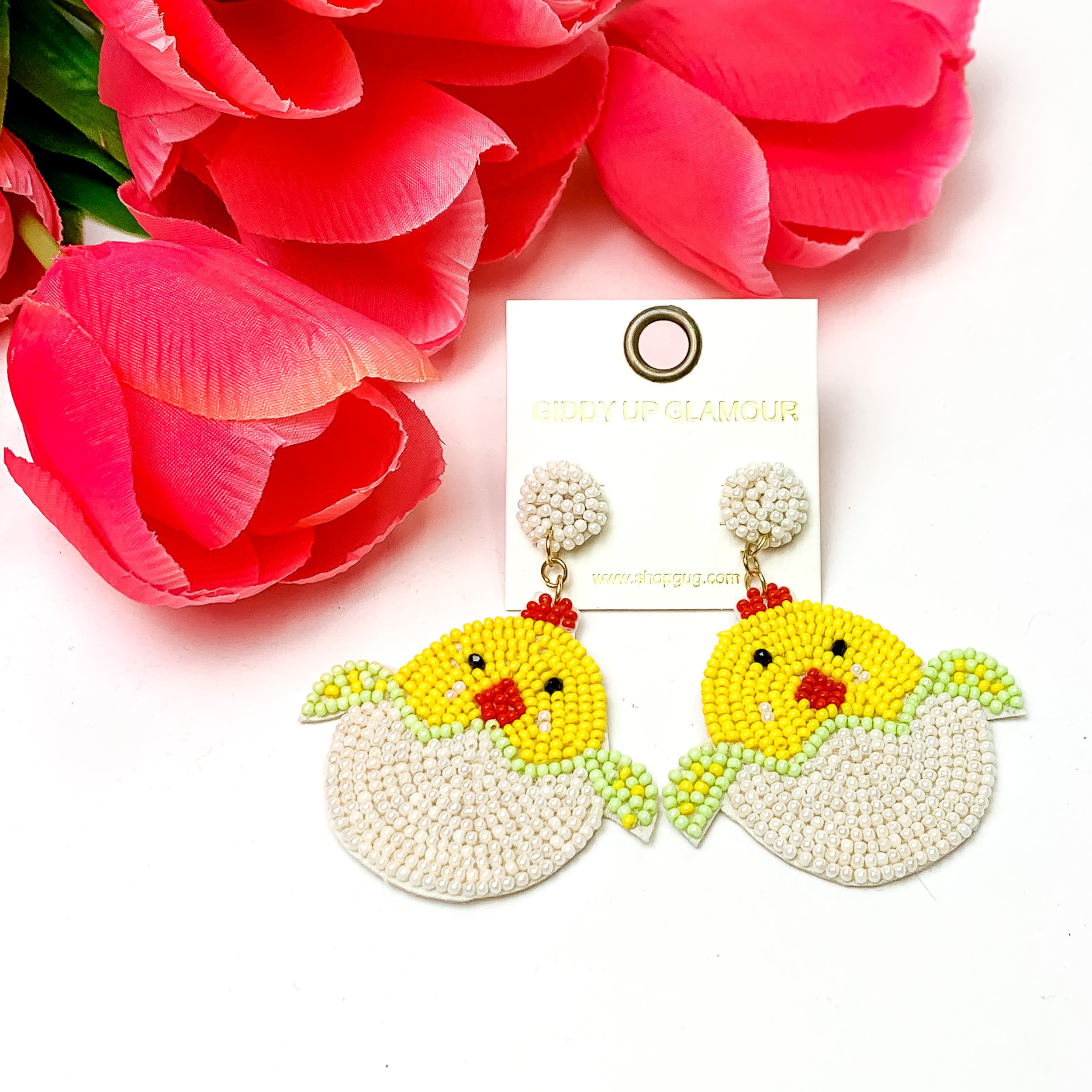 Beaded, white Easter egg earrings with a yellow beaded chick hatching. Pictured on white background with red-coral colored flowers.