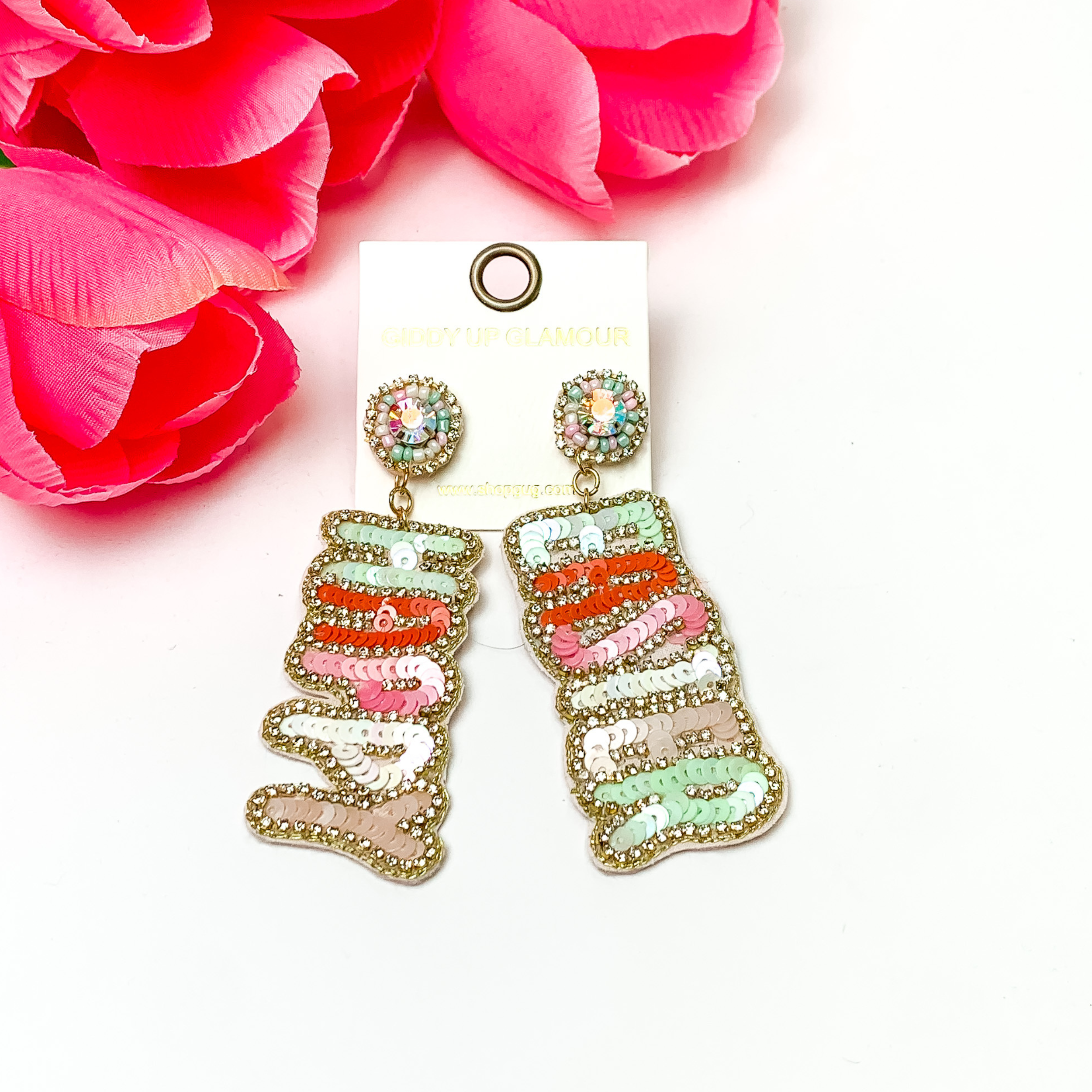 Pastel colored sequin beaded earrings. One earrings has the word "HAPPY" and the other earring has the word "EASTER" and each letter is a different pastel color. The words are hanging on clear crystal studs with nuetral colored beads around the crystal. These earrings are pictured on a white background with red- coral flowers in the left corner.