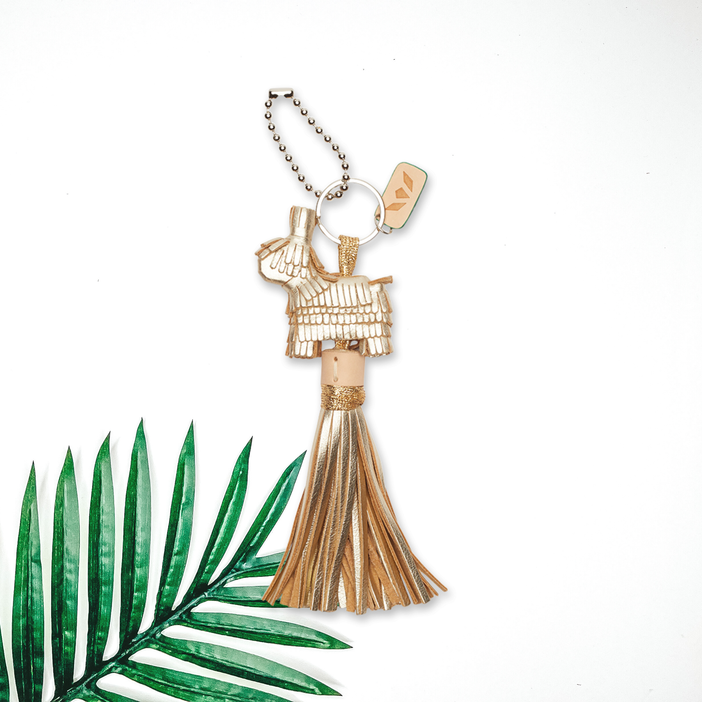 Centered in the middle of the picture is a gold donkey charm with gold tassels. To the right of the charm is a palm leaf, all on a white background. 