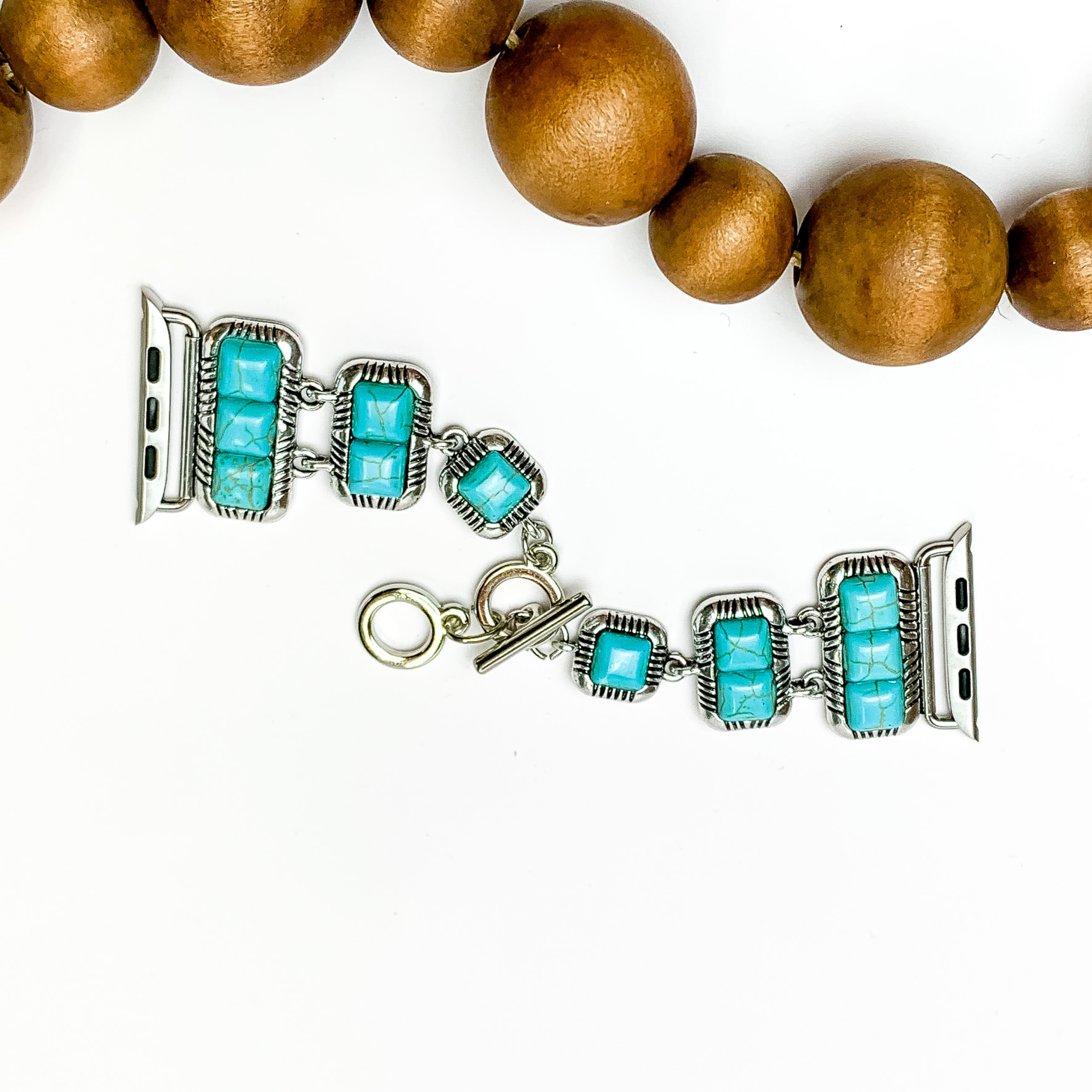 Silver smart watch band with five turquoise stones in each side with a toggle clasp. Turquoise stones are in square shape going from three stones to two stones and then one stone right before the toggle clasp. the watch is pictured on a white background with wooden beads above the watch.