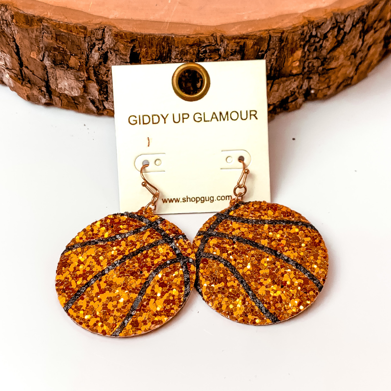 Pictured are circle drop glitter orange basketball earrings. They are propped up on a wooden half circle on a white background.