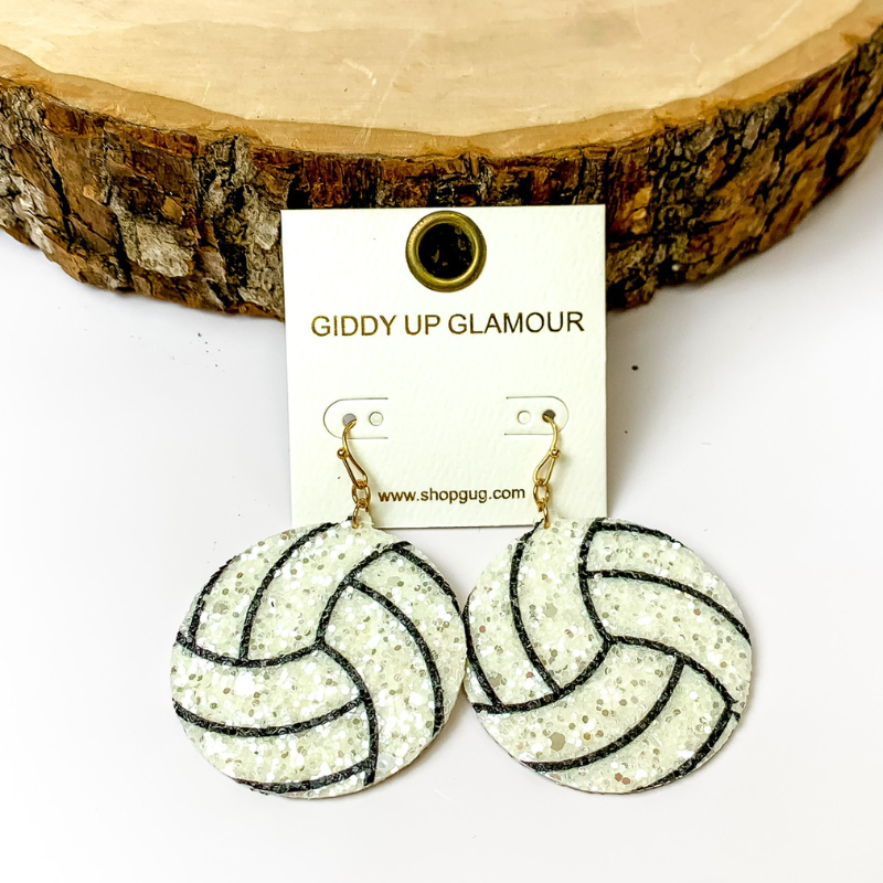 Pictured is circle drop glitter volleyball earrings in white. They are propped up on a circle piece of wood on a white background.